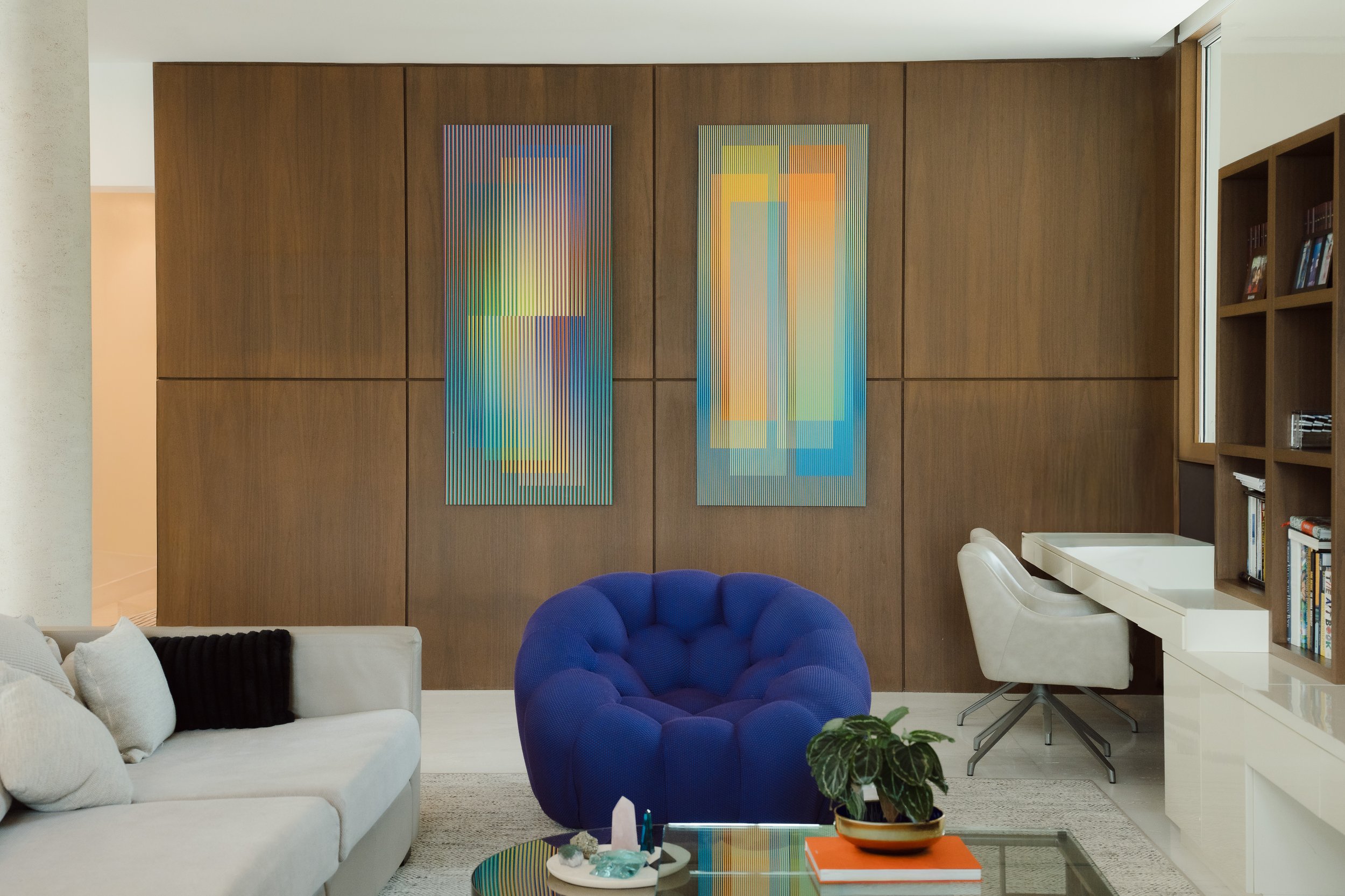 Living room with a white sofa, a blue cushioned chair, a white desk with a beige chair, wooden wall panels with colorful abstract art, and a bookshelf on the right side.