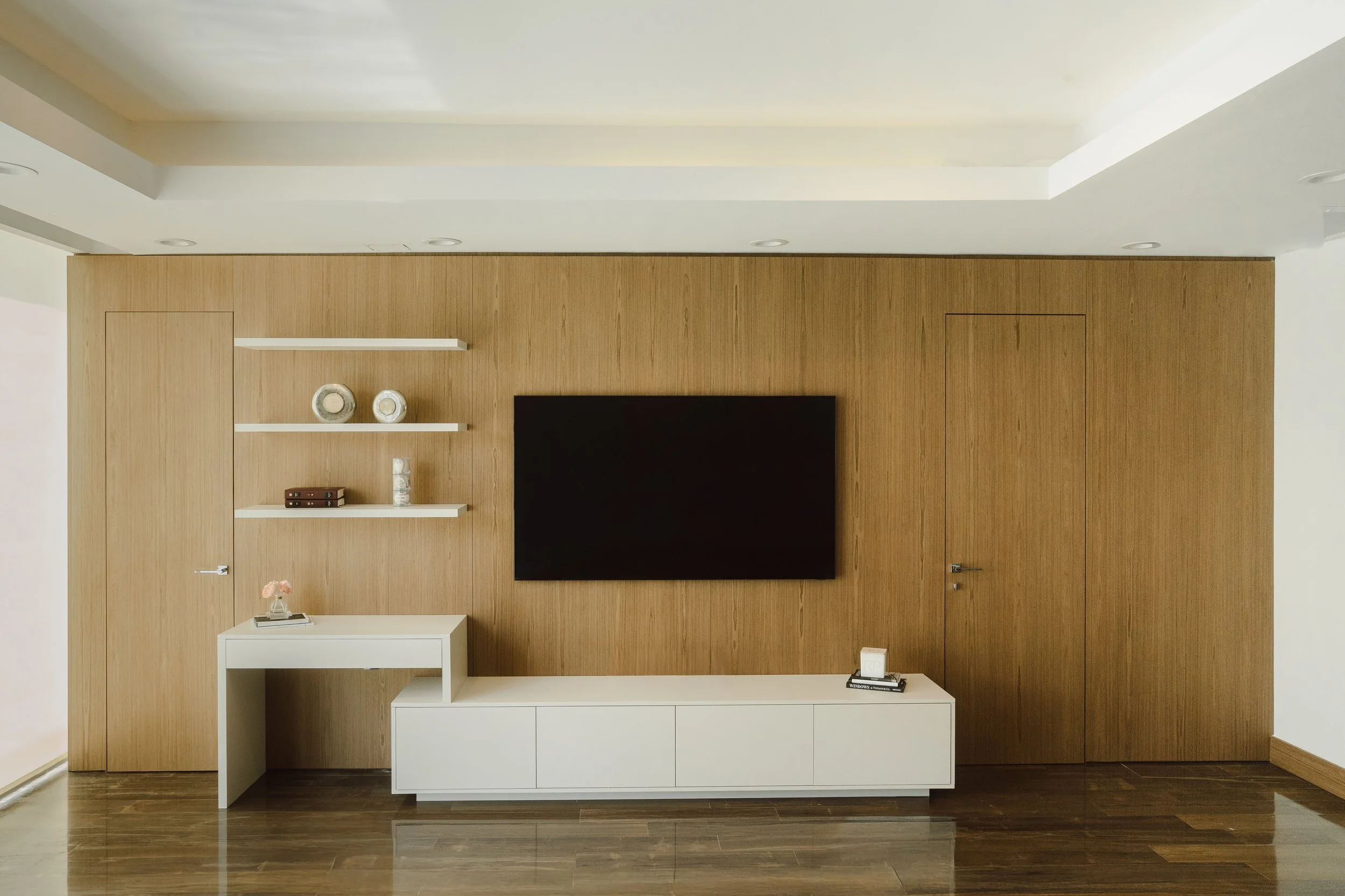 Modern living room wall with wooden paneling, mounted flat-screen TV, white shelves with vases and books, white cabinet with decor, hardwood floor, and a door on the right.