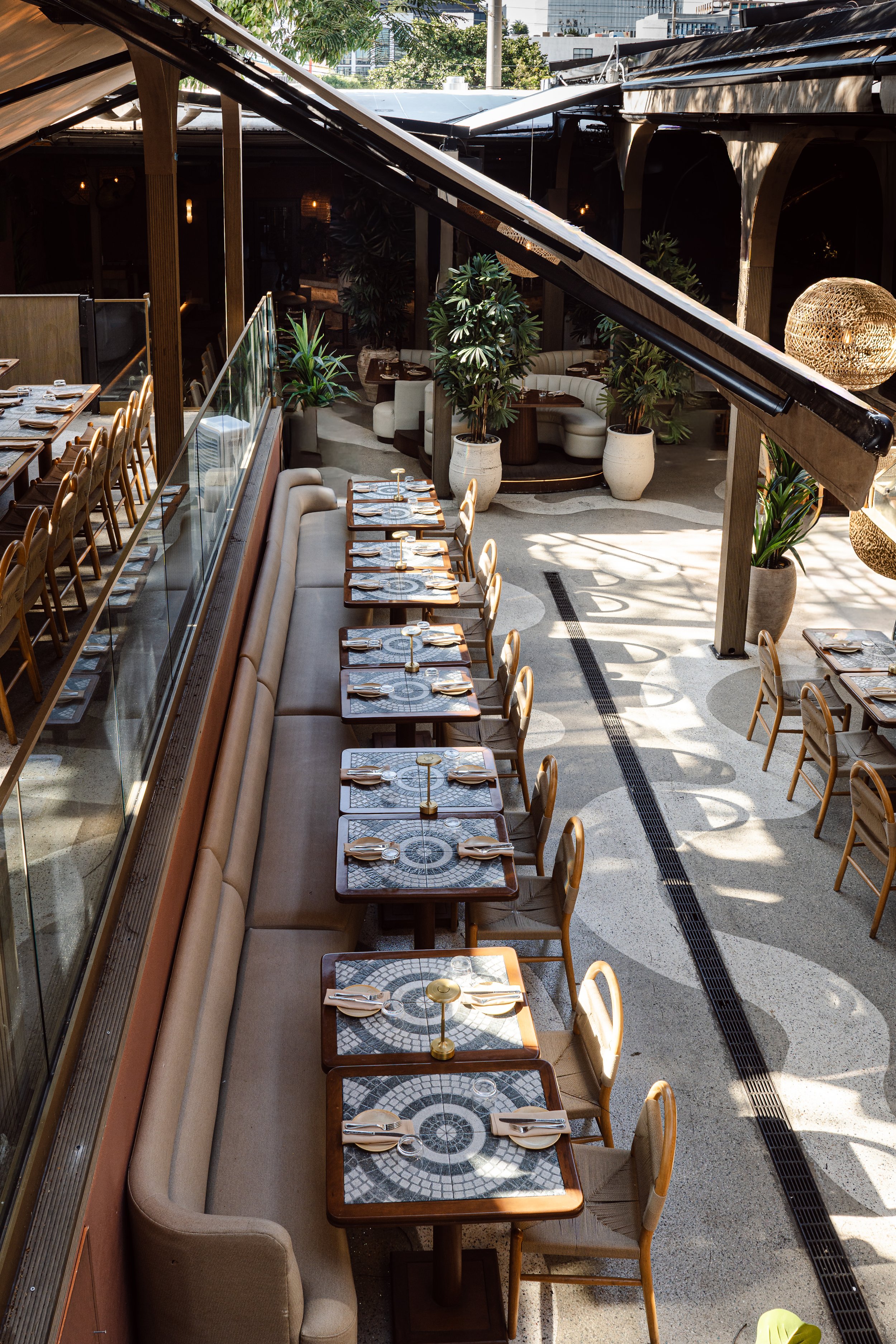 Empty indoor restaurant dining area with tables and chairs, decorated with potted plants, illuminated by sunlight.