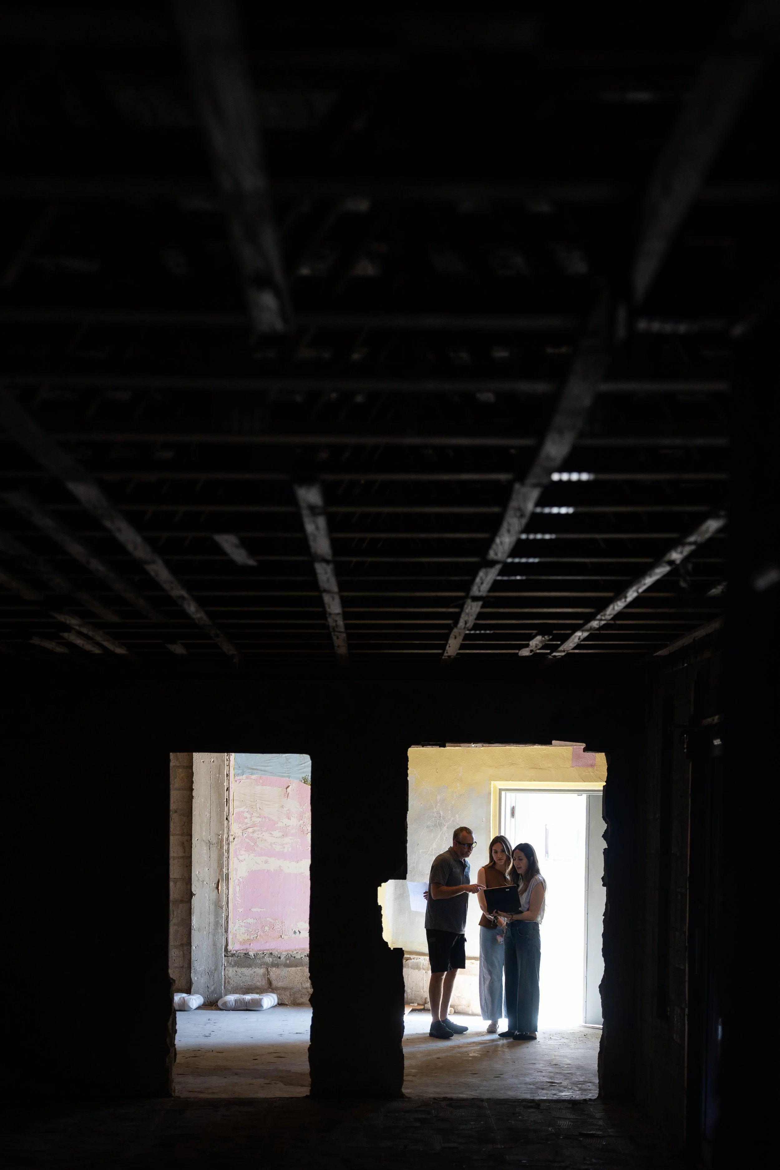 Three people standing in a room with unfinished walls, browsing a tablet or phone, with a large open window in the background.