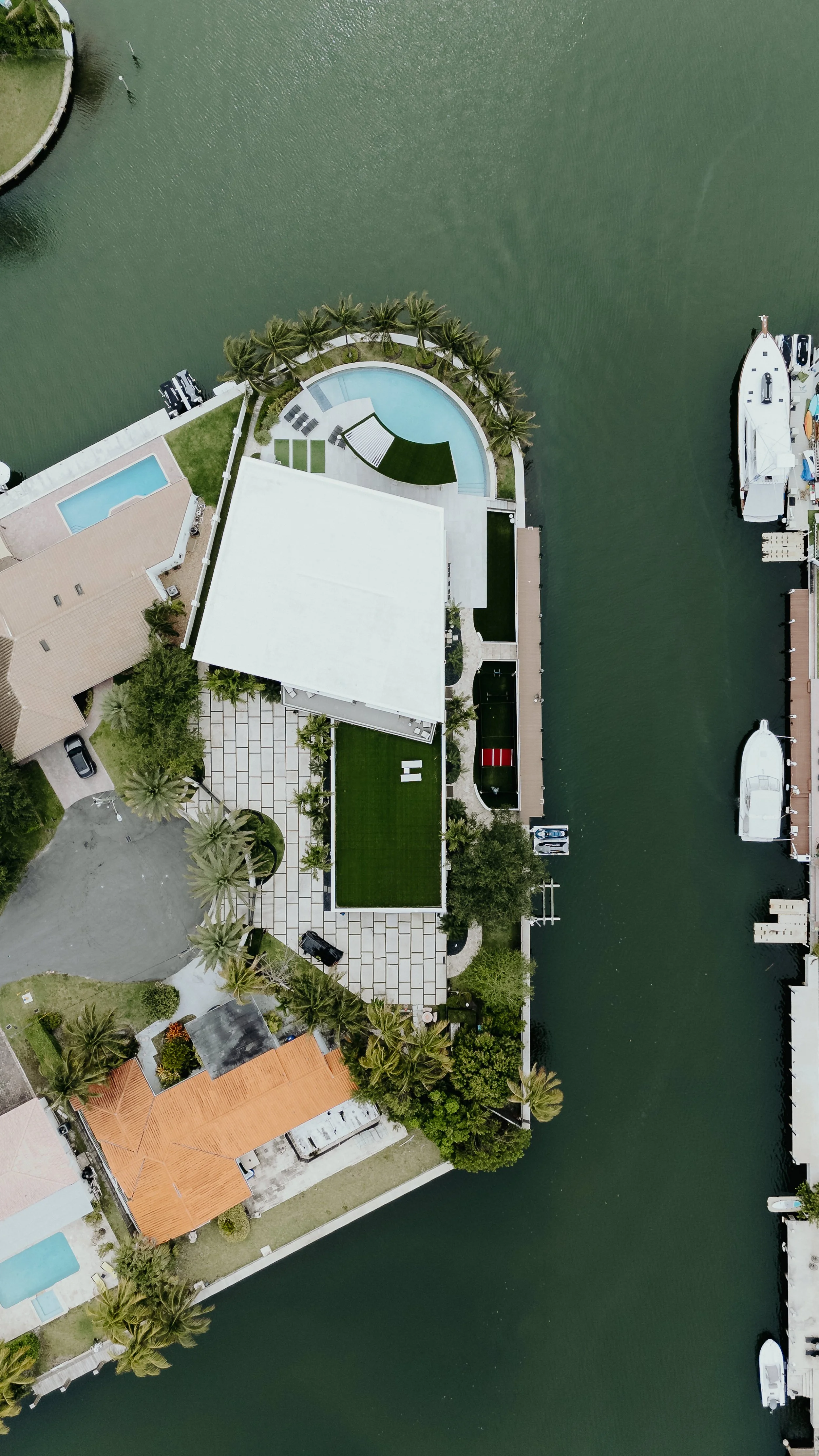 Aerial view of a waterfront property with a house, swimming pool, palm trees, and boats docked along the waterway.