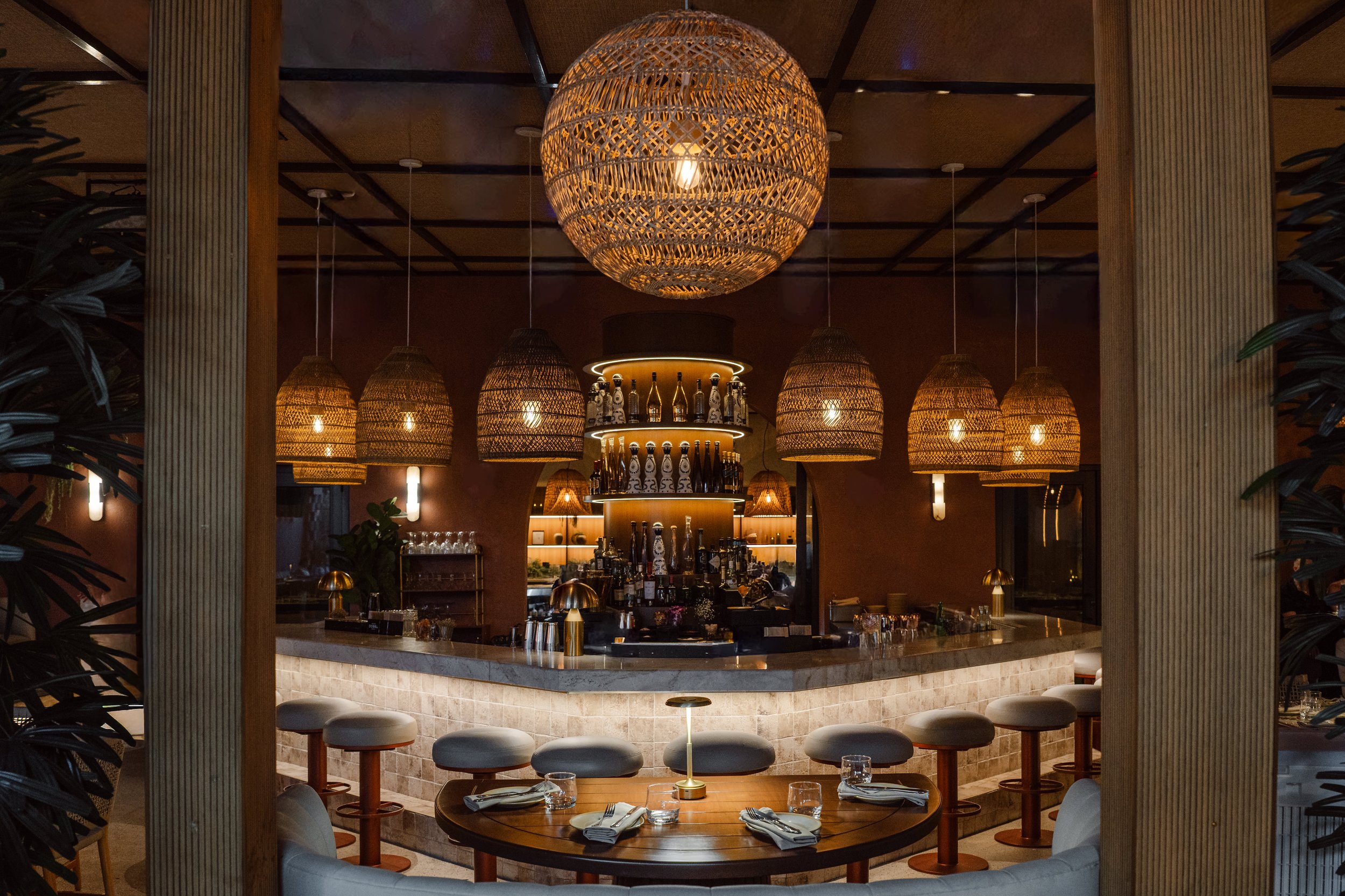 Interior of a restaurant or bar with a central bar counter, hanging wicker light fixtures, and bar shelves with bottles, illuminated by warm lighting.