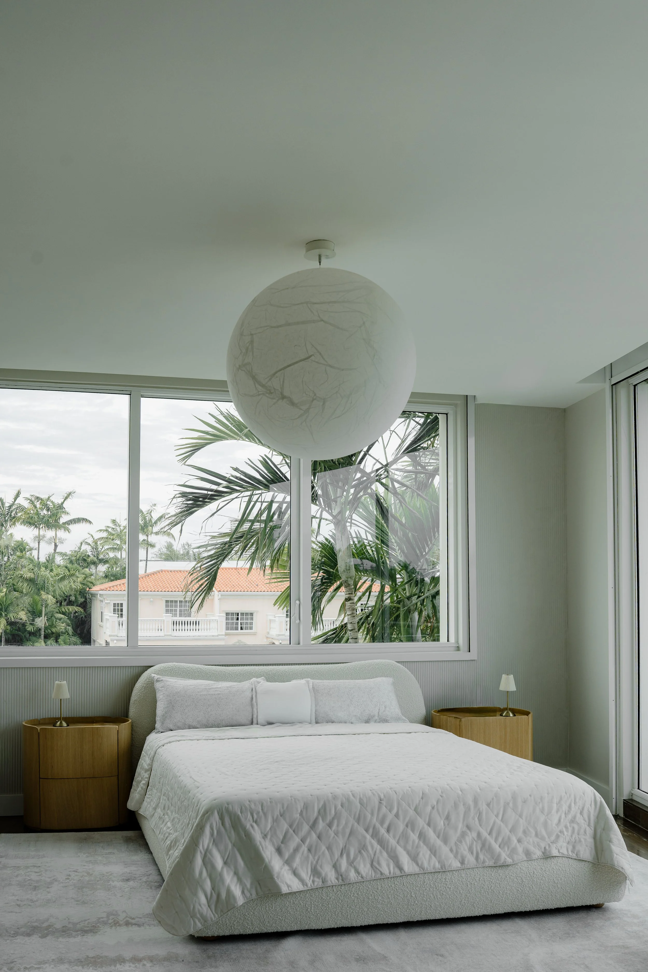 A bedroom with a large window showing a view of palm trees and houses outside. There is a white bed with a quilted bedspread, two wooden nightstands with small lamps, and a large spherical paper lantern hanging from the ceiling.