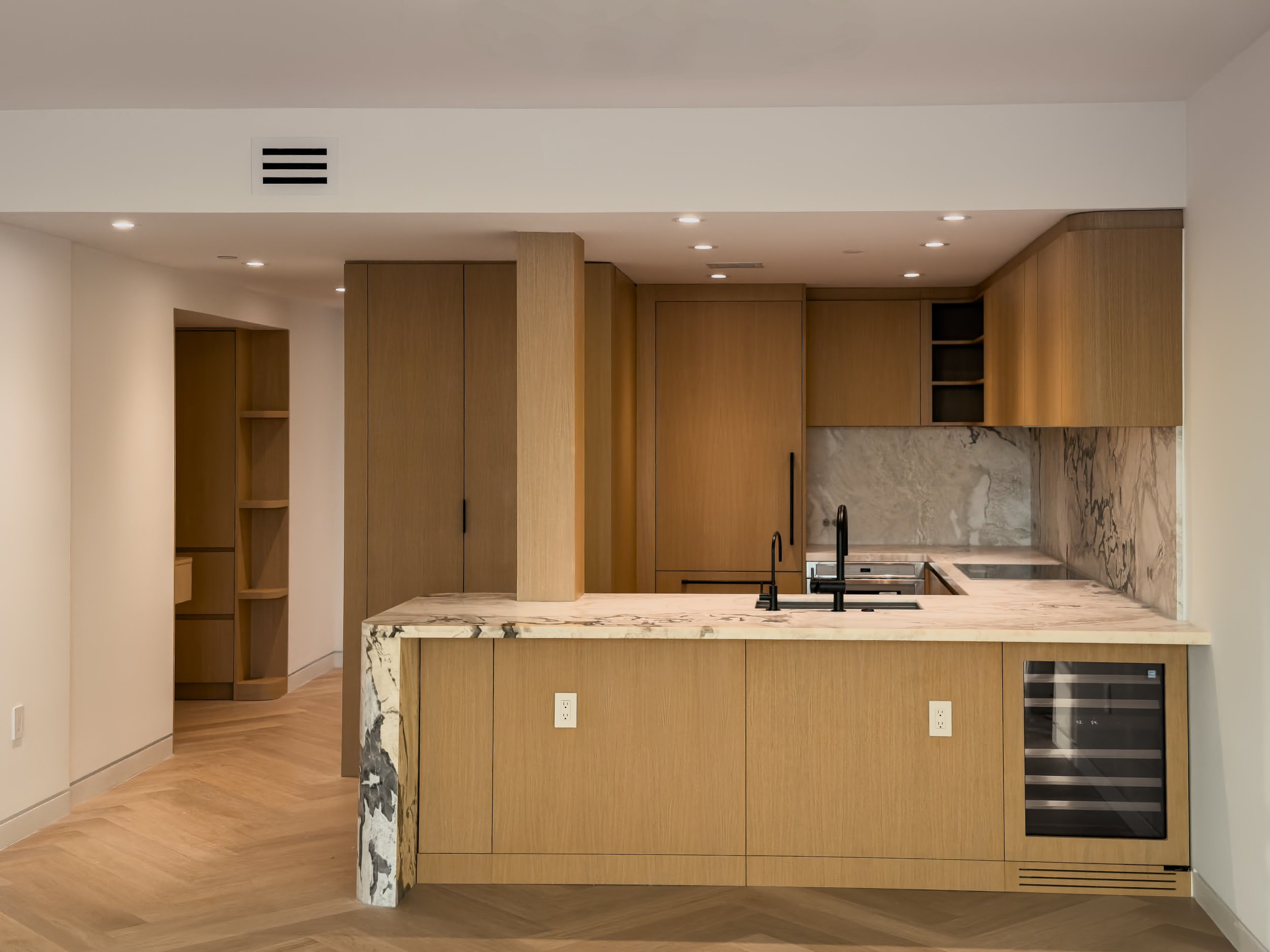 Modern kitchen with wooden cabinets, a marble island with a black sink and faucet, and a marble backsplash. Recessed ceiling lights and an electric oven are visible.
