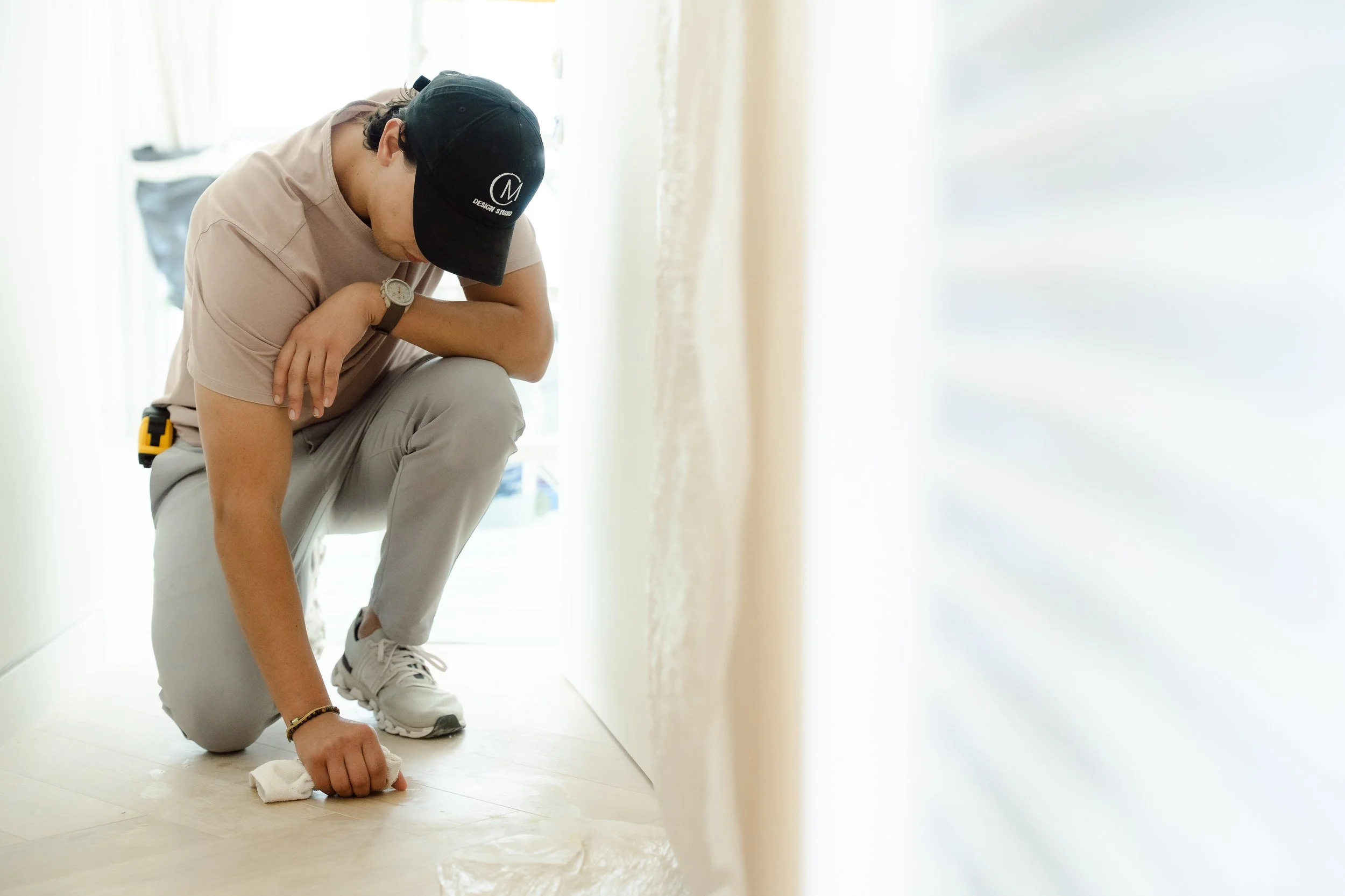 Person wearing a black cap and beige shirt cleaning a white floor with a cloth, with a bright background and light-colored walls.