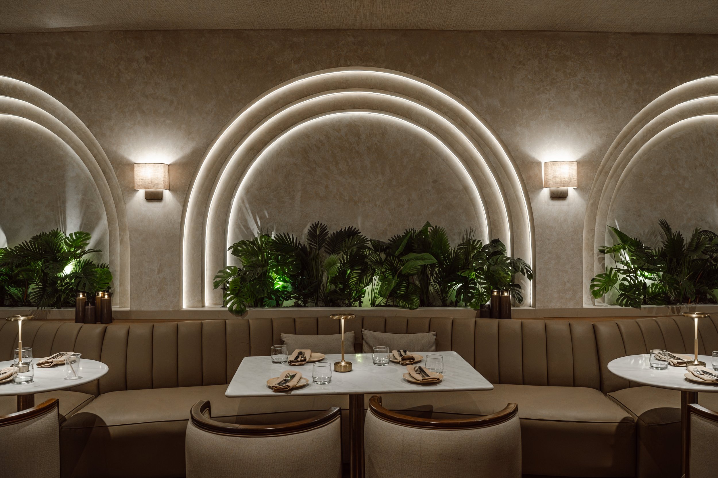 Indoor restaurant with a curved beige upholstered banquette, leafy green plants, wall-mounted light fixtures, and a decorative arch illuminated with lighting.