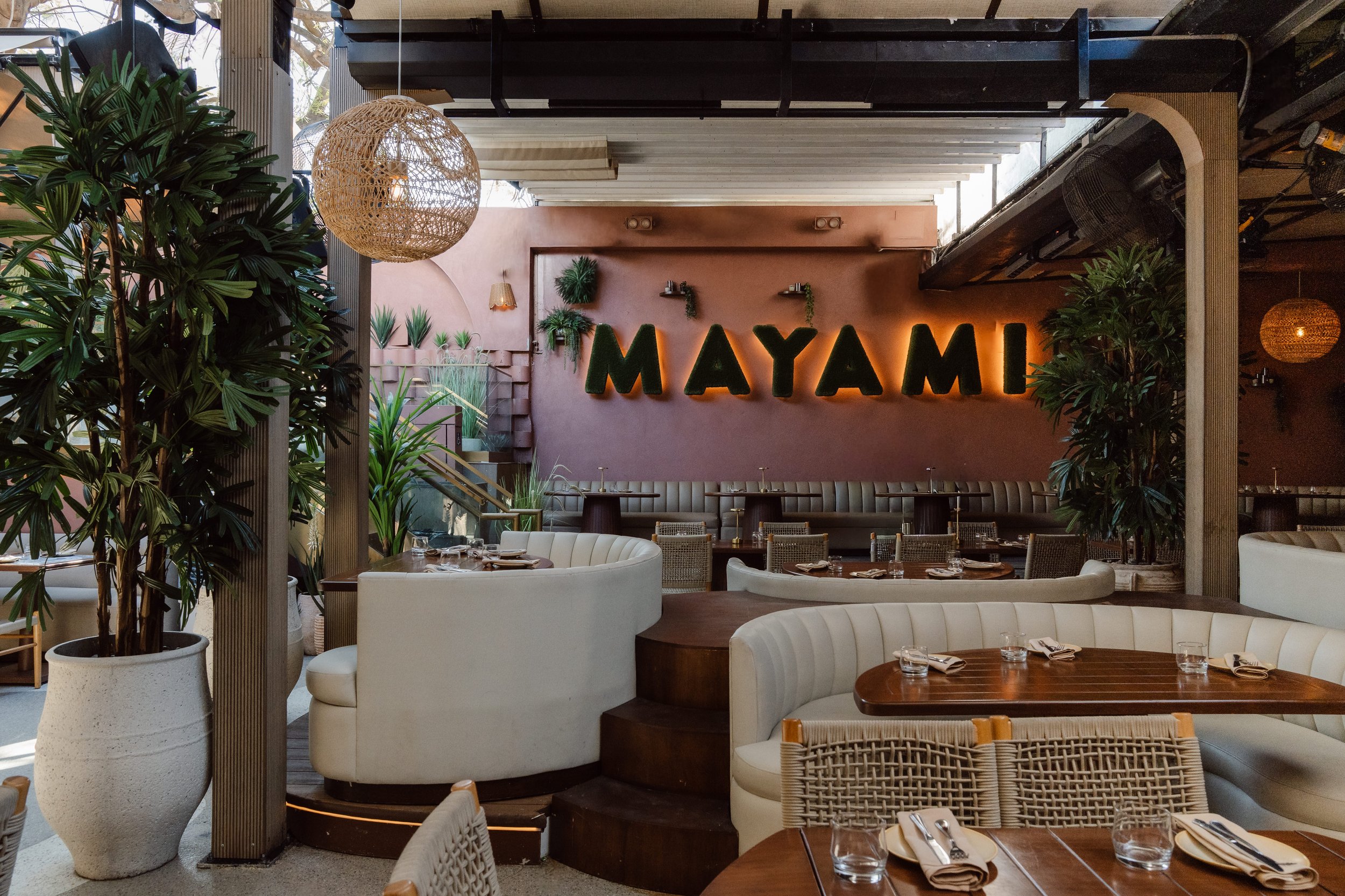 Indoor restaurant with modern decor, lush green plants, a large illuminated sign that says 'MAYAMI,' wicker pendant light, and tables set for dining.