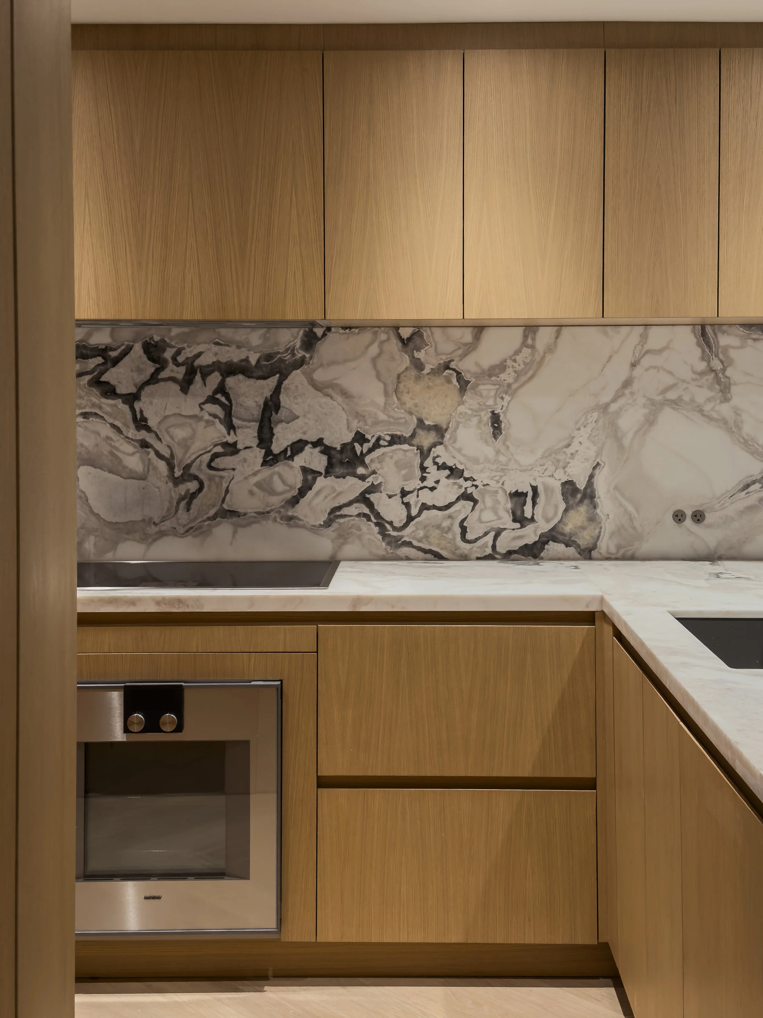 Modern kitchen with light wood cabinets, marble backsplash, and a built-in oven.