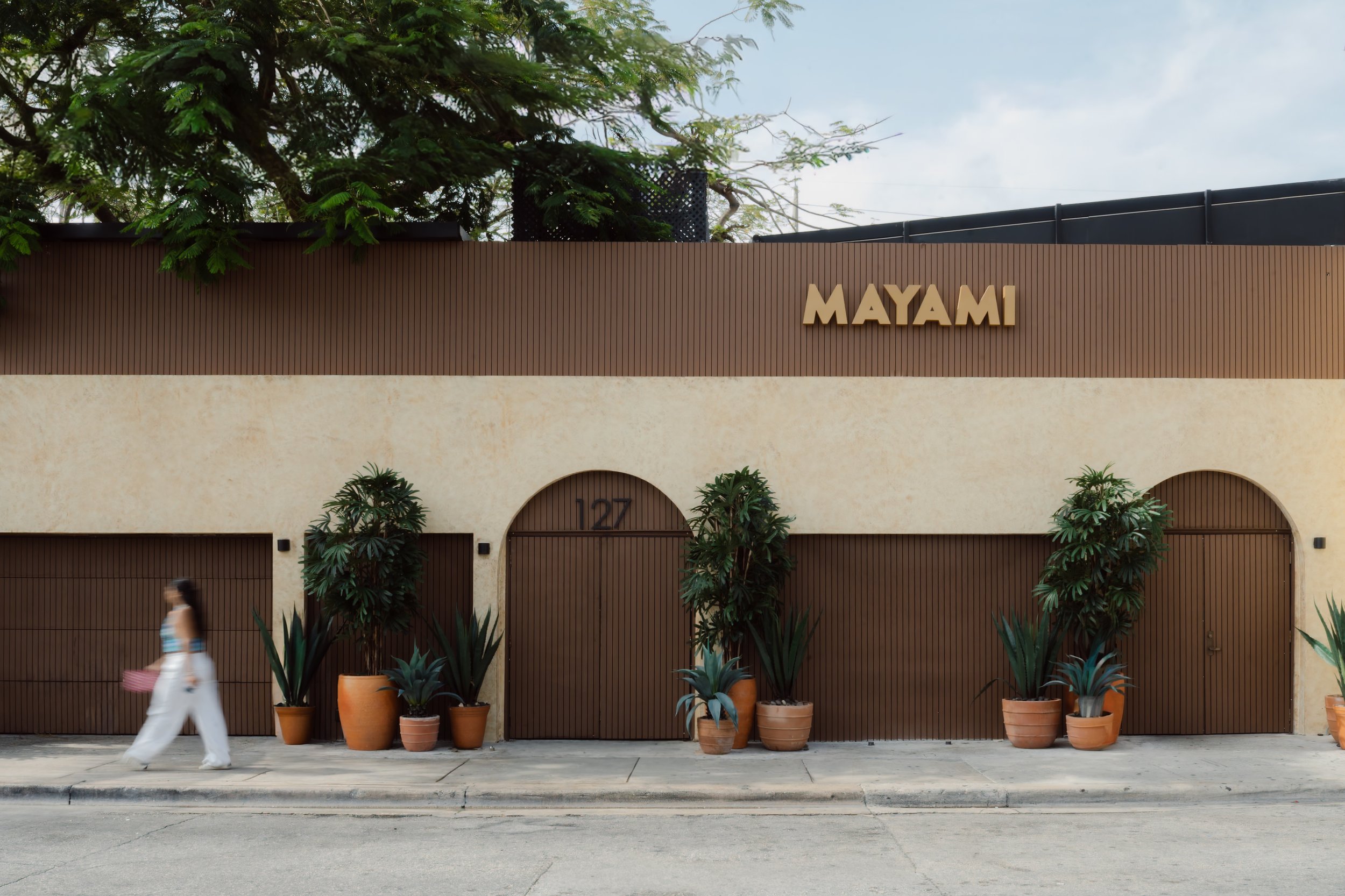 Exterior of a building with the name 'MAYAMI' on a brown wooden panel. Potted plants are lined up in front of the building, and a woman in white is walking past on the sidewalk. There are trees and a fence above the building.