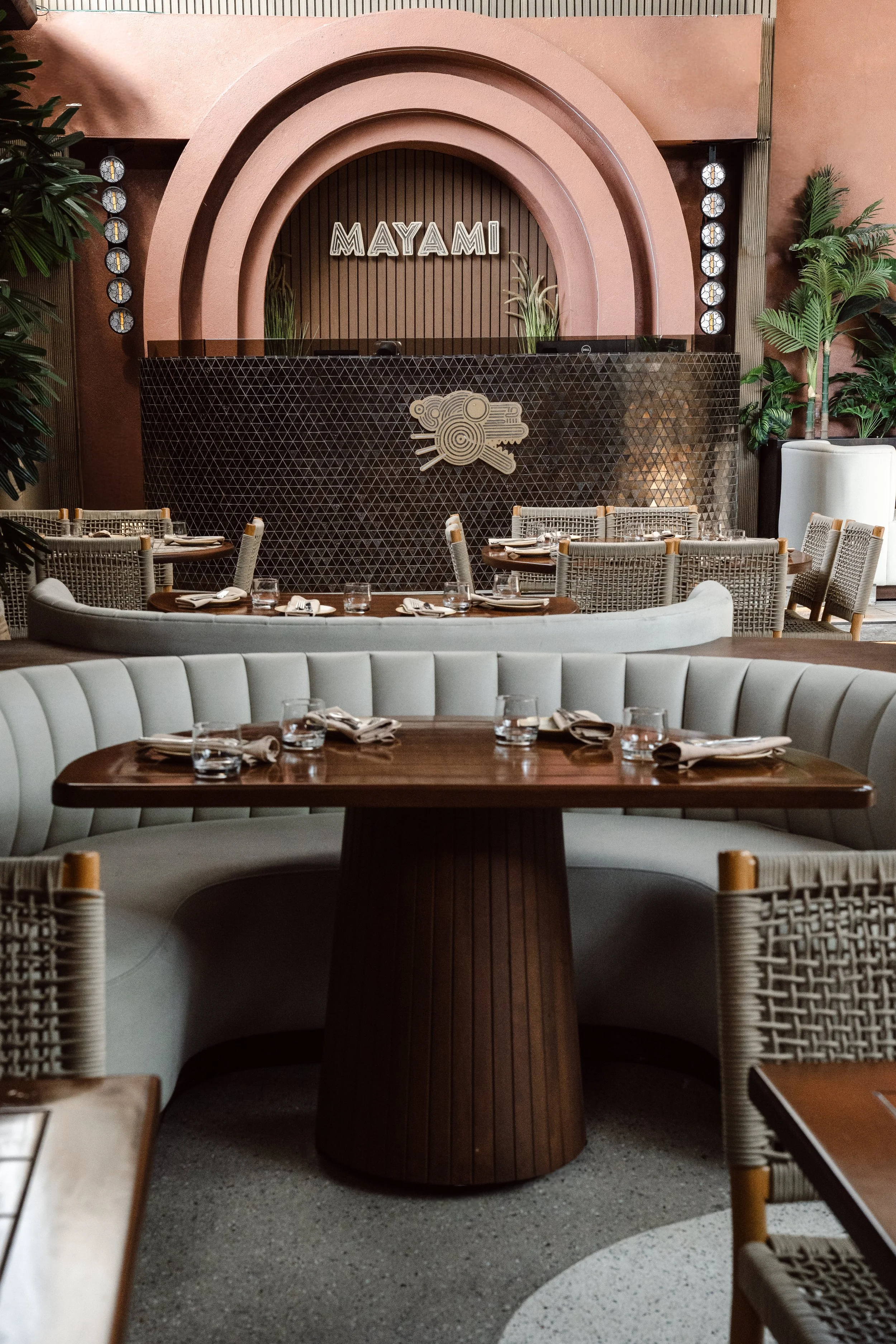 Interior of a restaurant with a curved wooden table, beige woven chairs, and a bar with a black geometric textured front. There are potted plants around and a sign that says 'MAYAMI' on the back wall, which is decorated with vertical wood paneling.