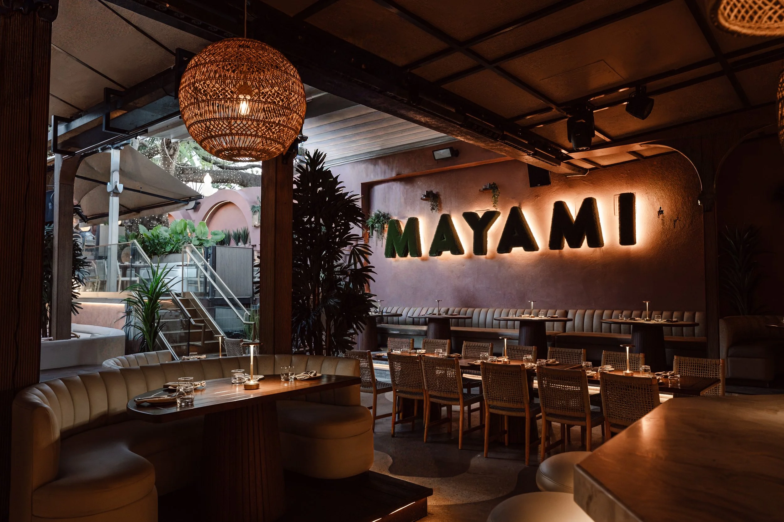 Interior of a stylish restaurant called Mayami, featuring a warm lighting ambiance, wooden furniture, a rattan hanging lamp, and lush green plants.