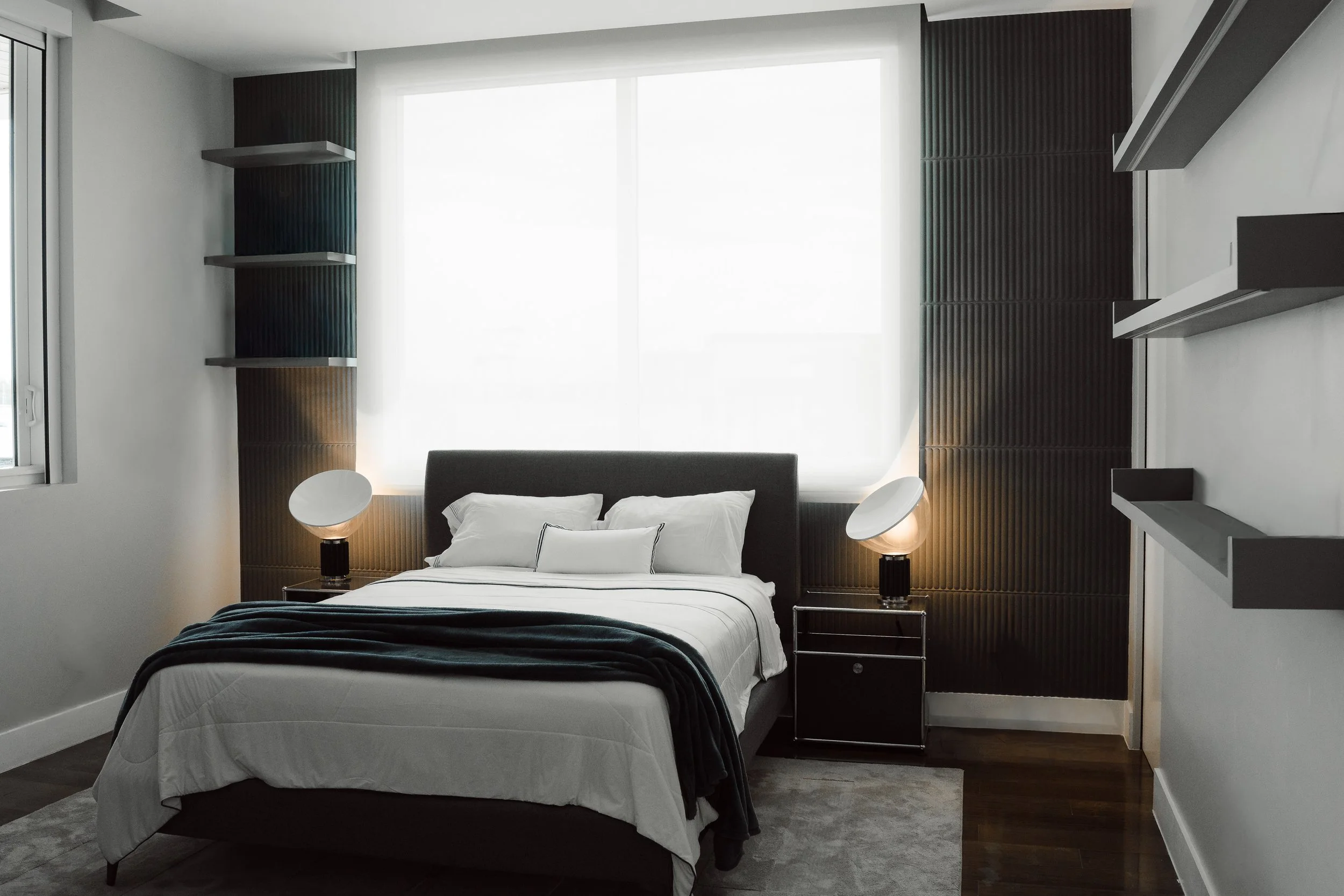 Modern bedroom with a large bed, white bedding, black and white pillows, two bedside tables with lamps, dark wood wall panels, and large window with white shades.