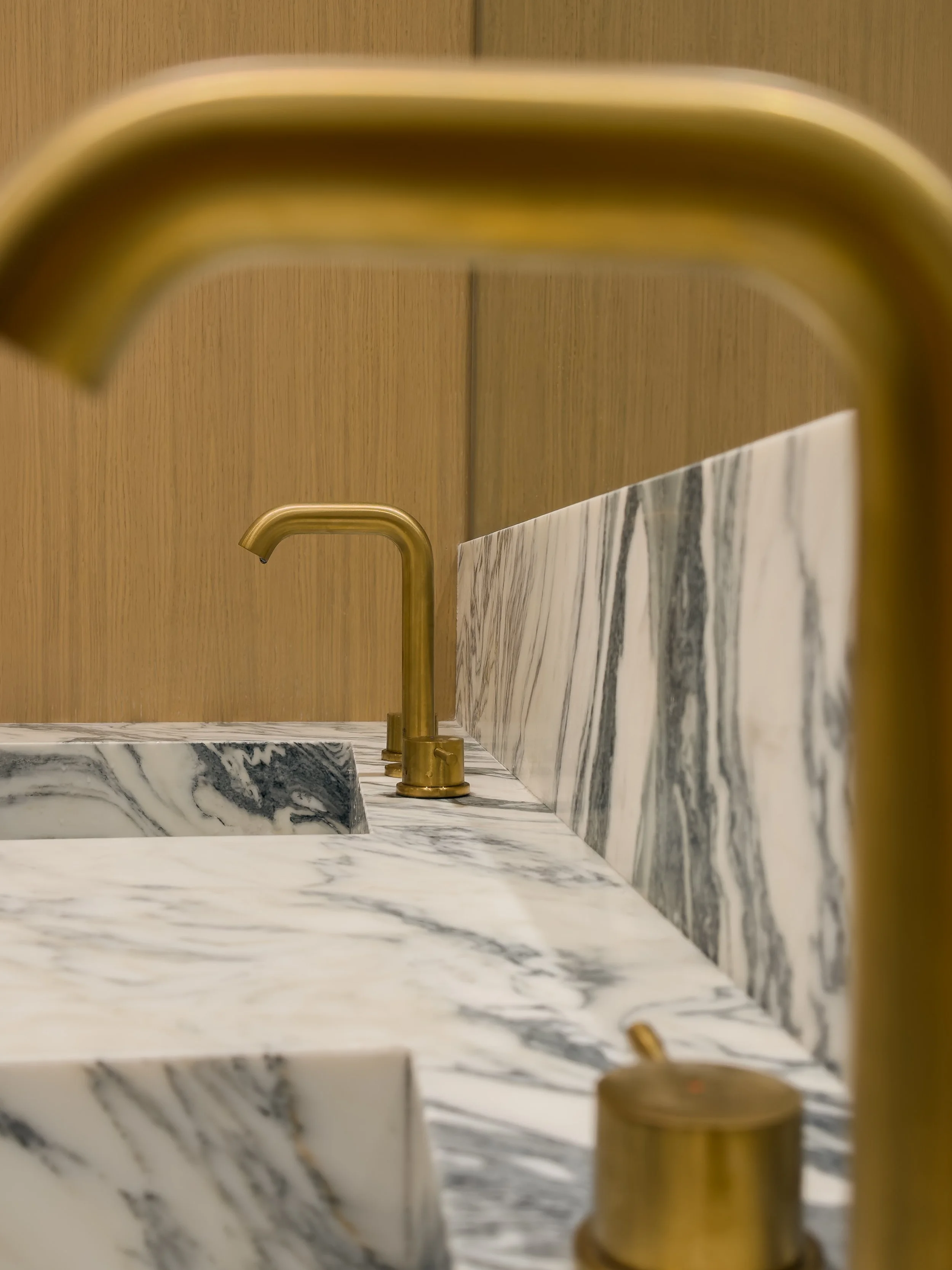 Close-up of a gold-colored faucet over a marble sink with a marble backsplash in a modern bathroom. The background features a wooden panel wall.