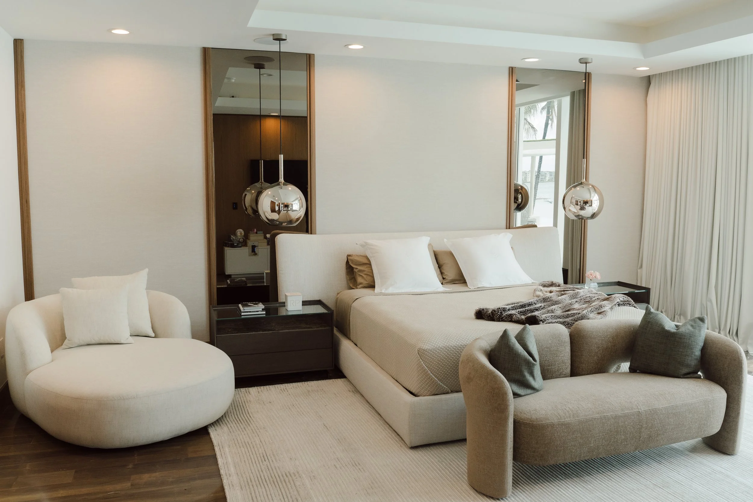 Modern hotel room with a large cushioned bed, a round white chair, and a side table. Mirrored panels and pendant lights hang above the bed. Large window with views of palm trees and the ocean.