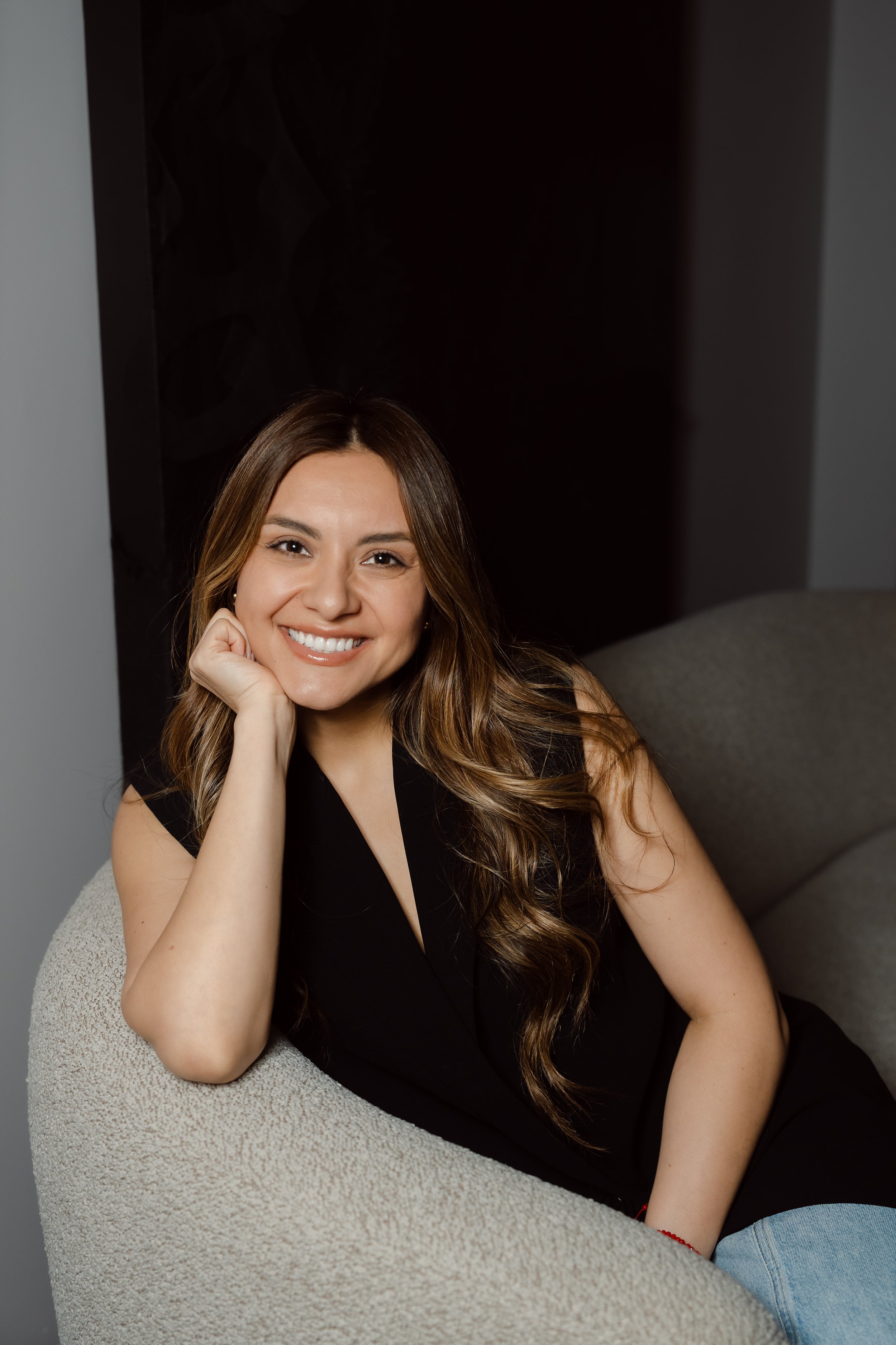 A young woman with long brown hair and a bright smile, resting her chin on her hand, sitting on a cushioned chair with a beige armrest. She is wearing a black top and blue jeans, with a gray and dark background.