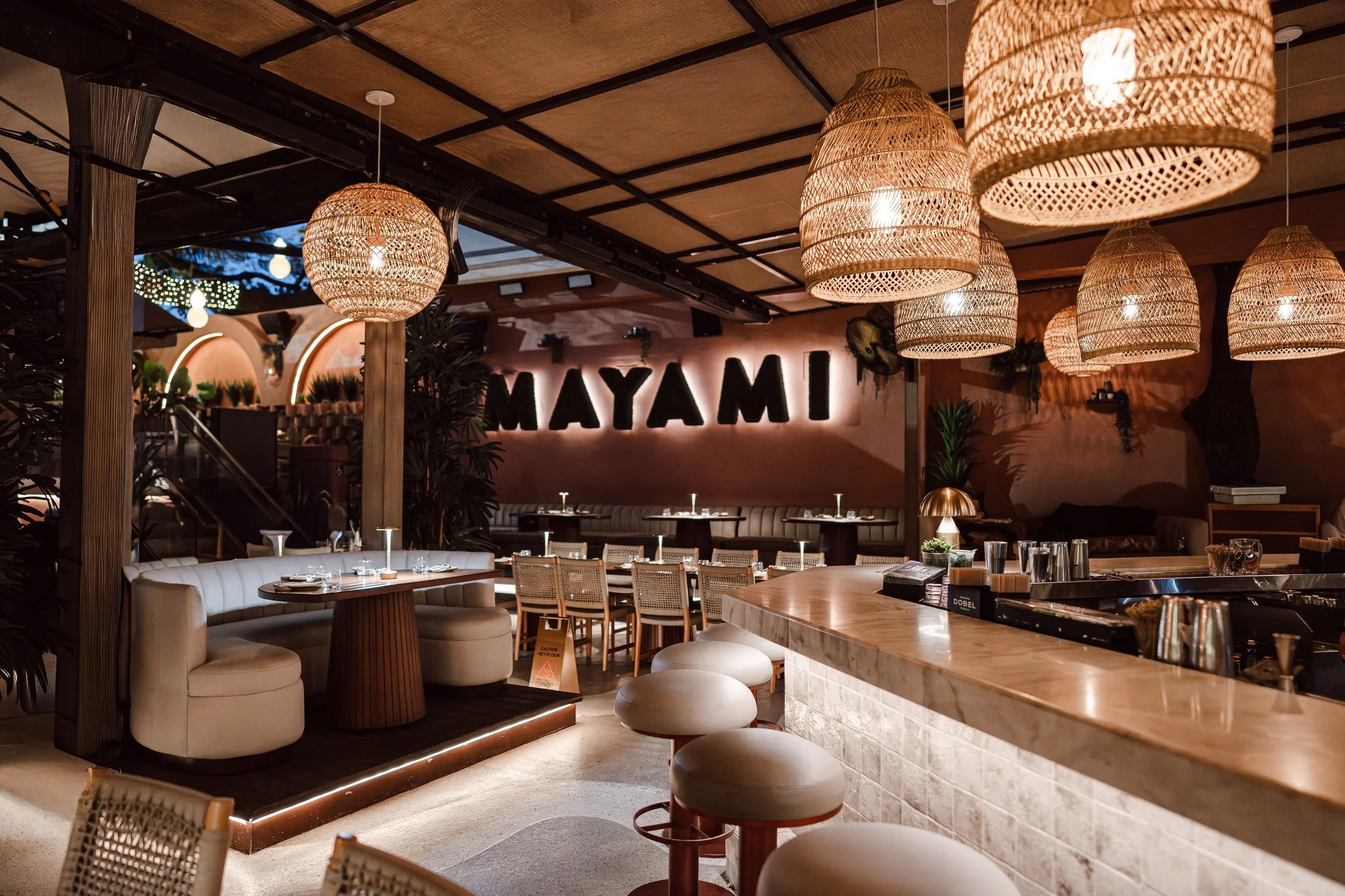Interior of a restaurant or bar with hanging wicker pendant lights, a curved booth seating area, tables with chairs, and a bar counter, with a sign in the background that says 'MAYA M'.