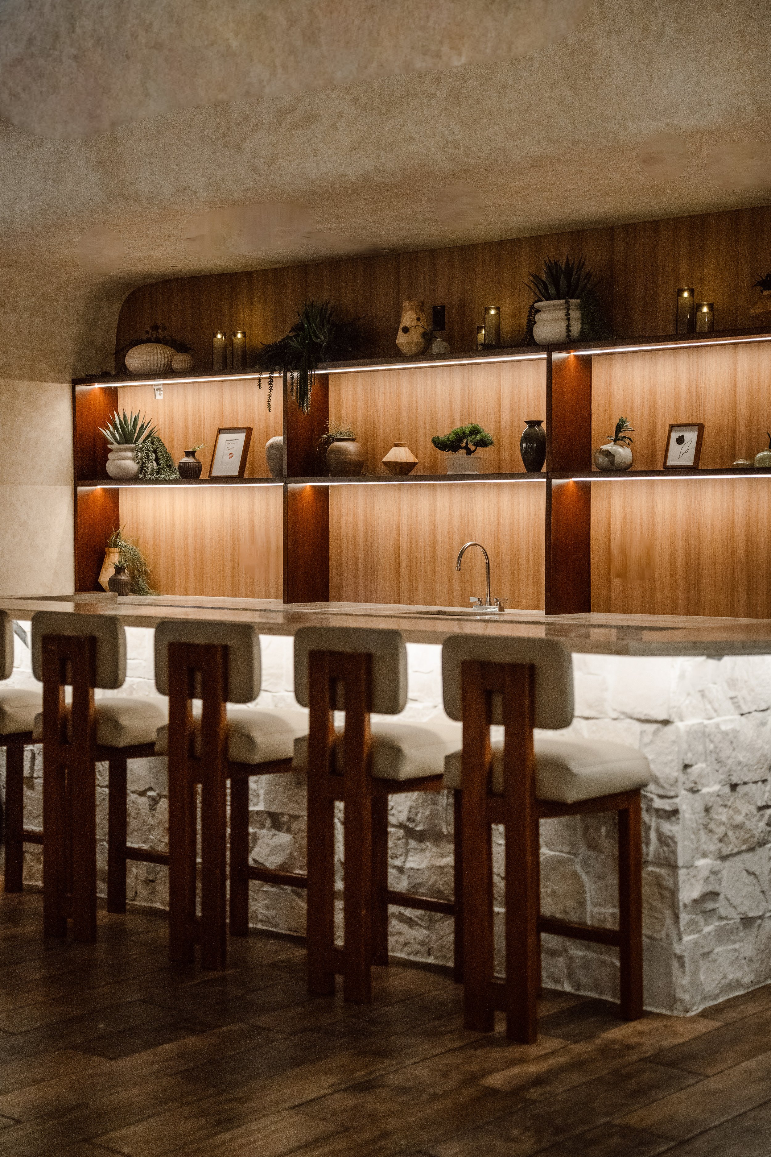 Wooden bar counter with high chairs, decorative vases, framed pictures, potted plants, and a stone wall below the counter, in a cozy, dimly lit cafe or restaurant.