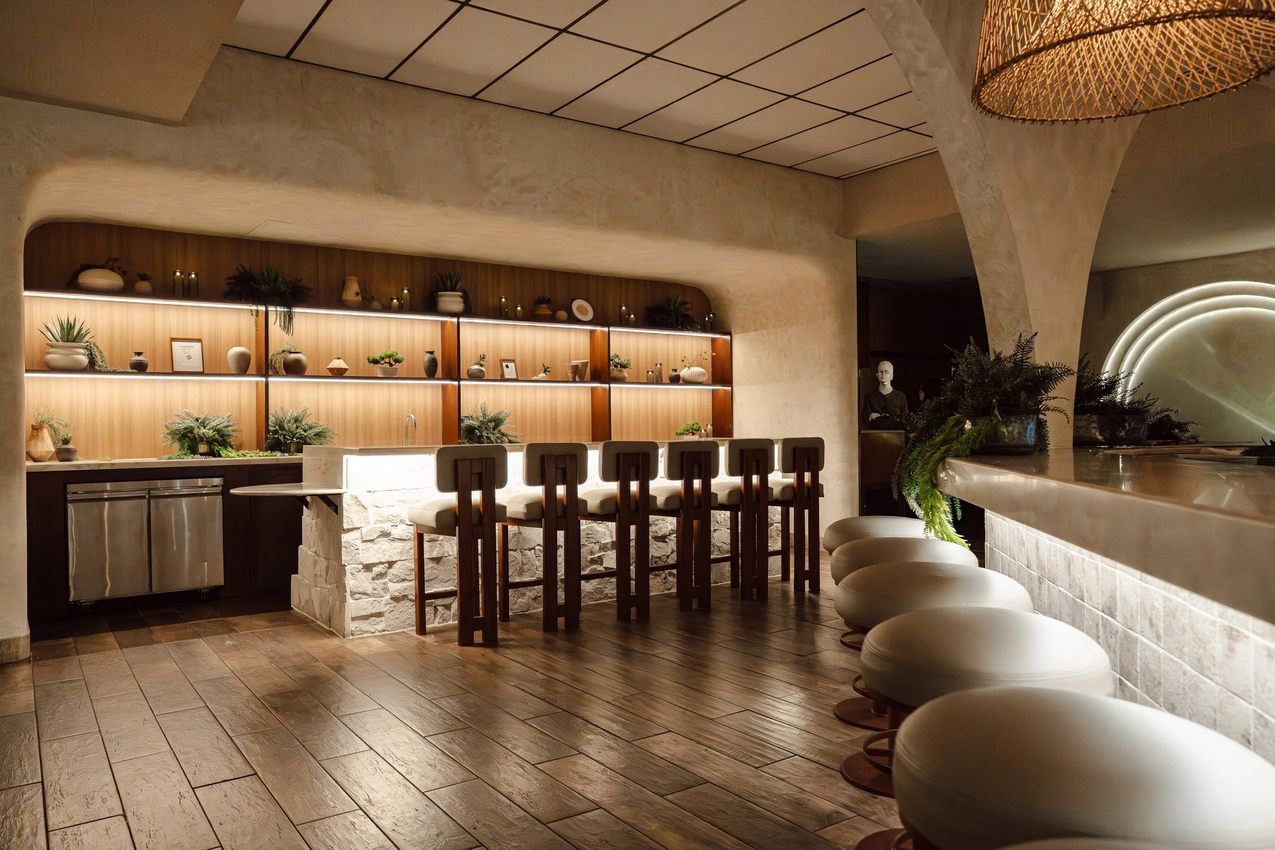 A bar area with a curved light-colored wall and wooden shelf displaying pottery and plants. Bar chairs are lined up along a counter with a row of round, white stools on the right. The ceiling is tiled, and there is warm lighting throughout the space,