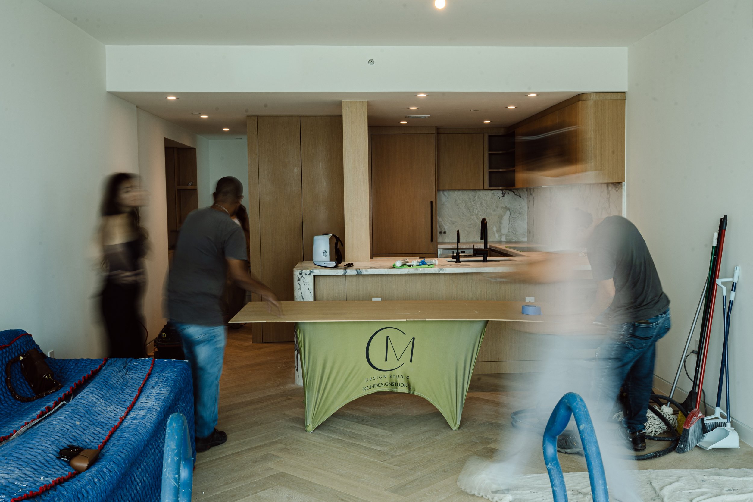 People working in a modern kitchen with wooden cabinets, marble backsplash, and a marble kitchen island. There is a blue sofa in the foreground and cleaning tools on the right side.