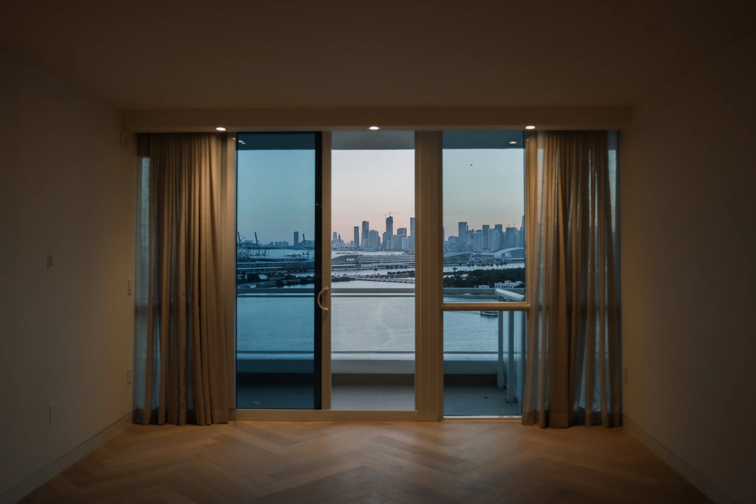Indoor room with large glass doors open to a balcony, showing a city skyline with skyscrapers and a body of water beyond.