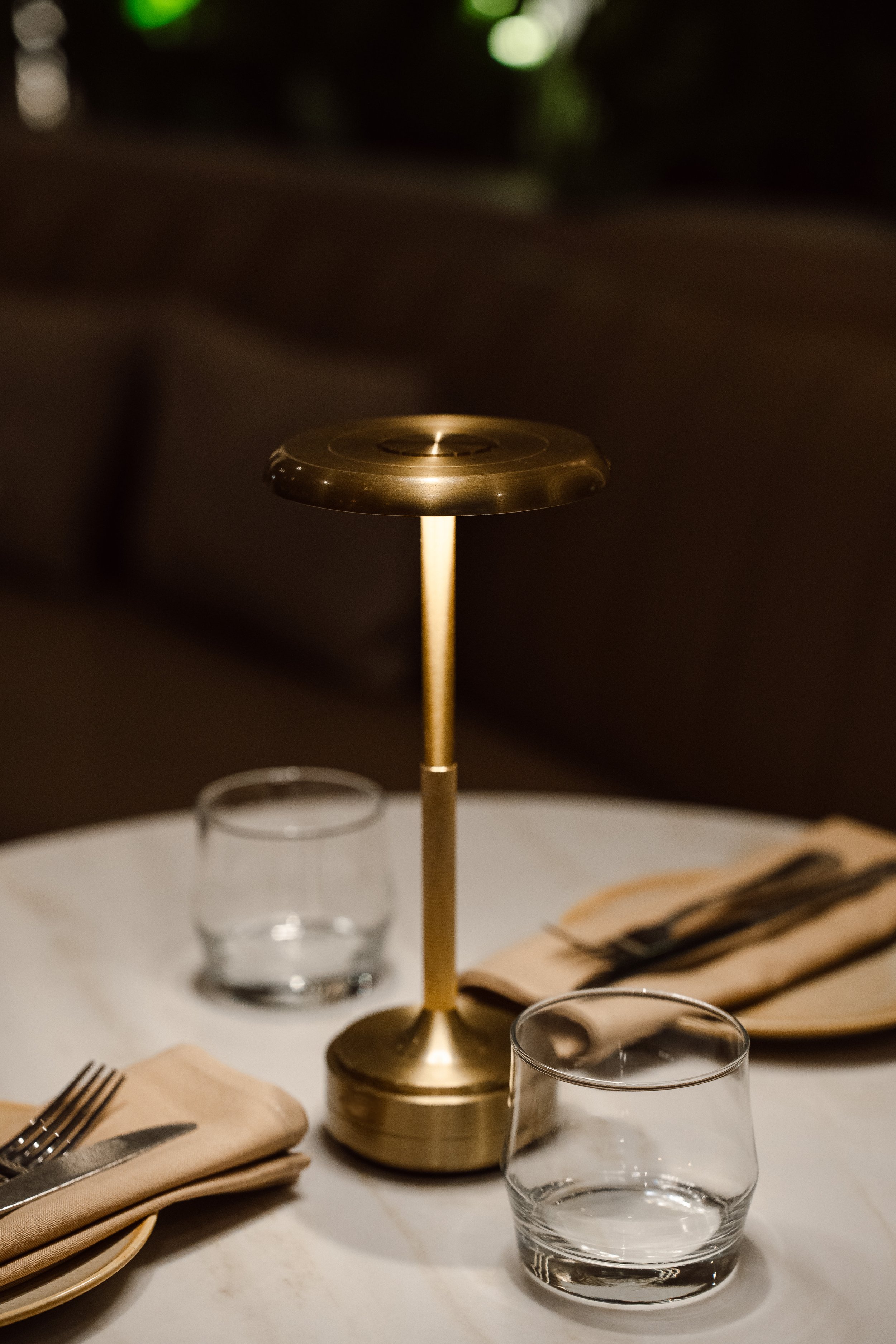 A dining table setup with a gold-colored centerpiece, clear glass cups, and beige napkins with silverware. The background is dark and out of focus.