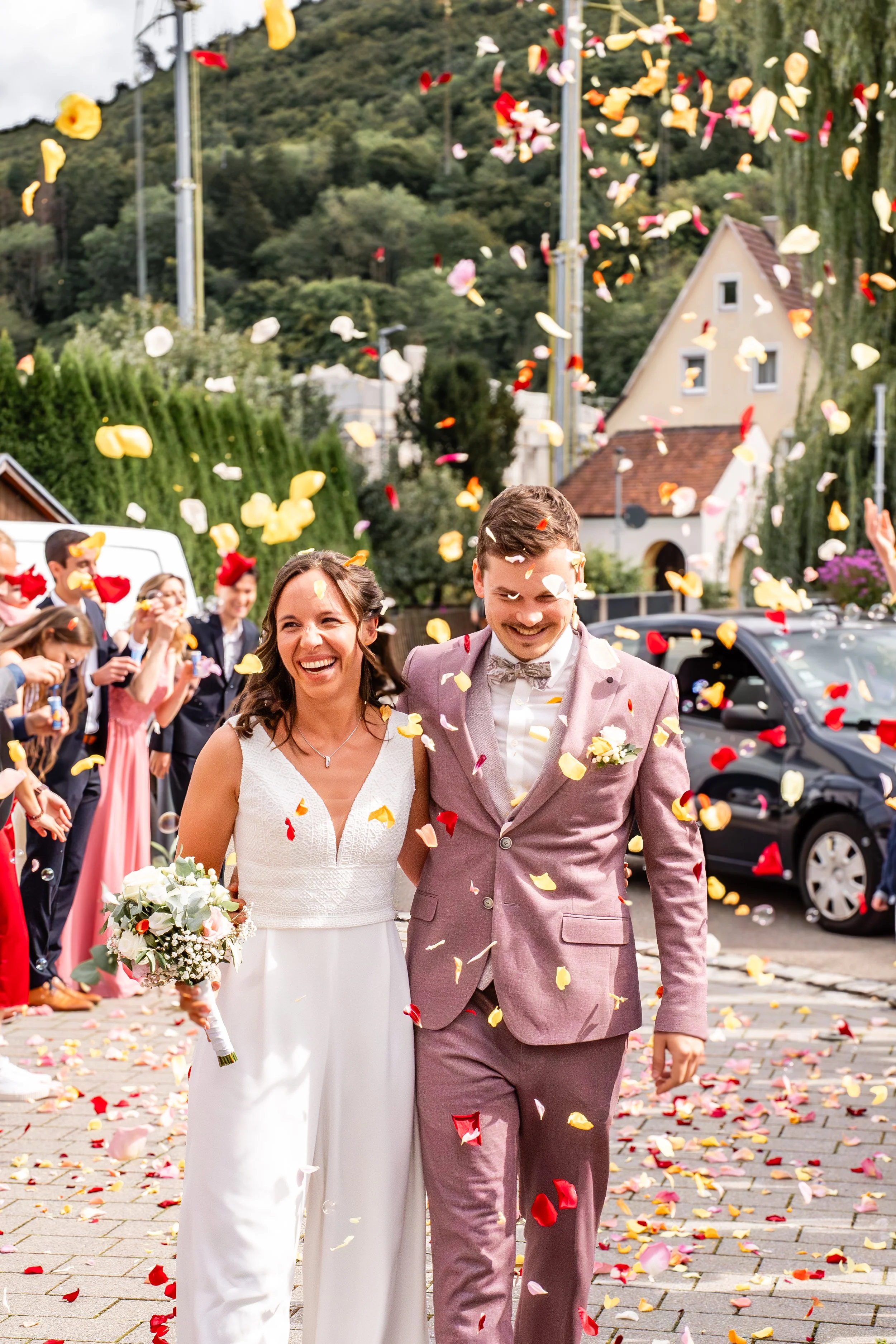 Ein glückliches Brautpaar läuft Hand in Hand durch eine confetti-übersäte Straße während einer Hochzeitsfeier, umgeben von Freunden und Familie. Im Hintergrund sind Häuser und grüne Hügel sichtbar.