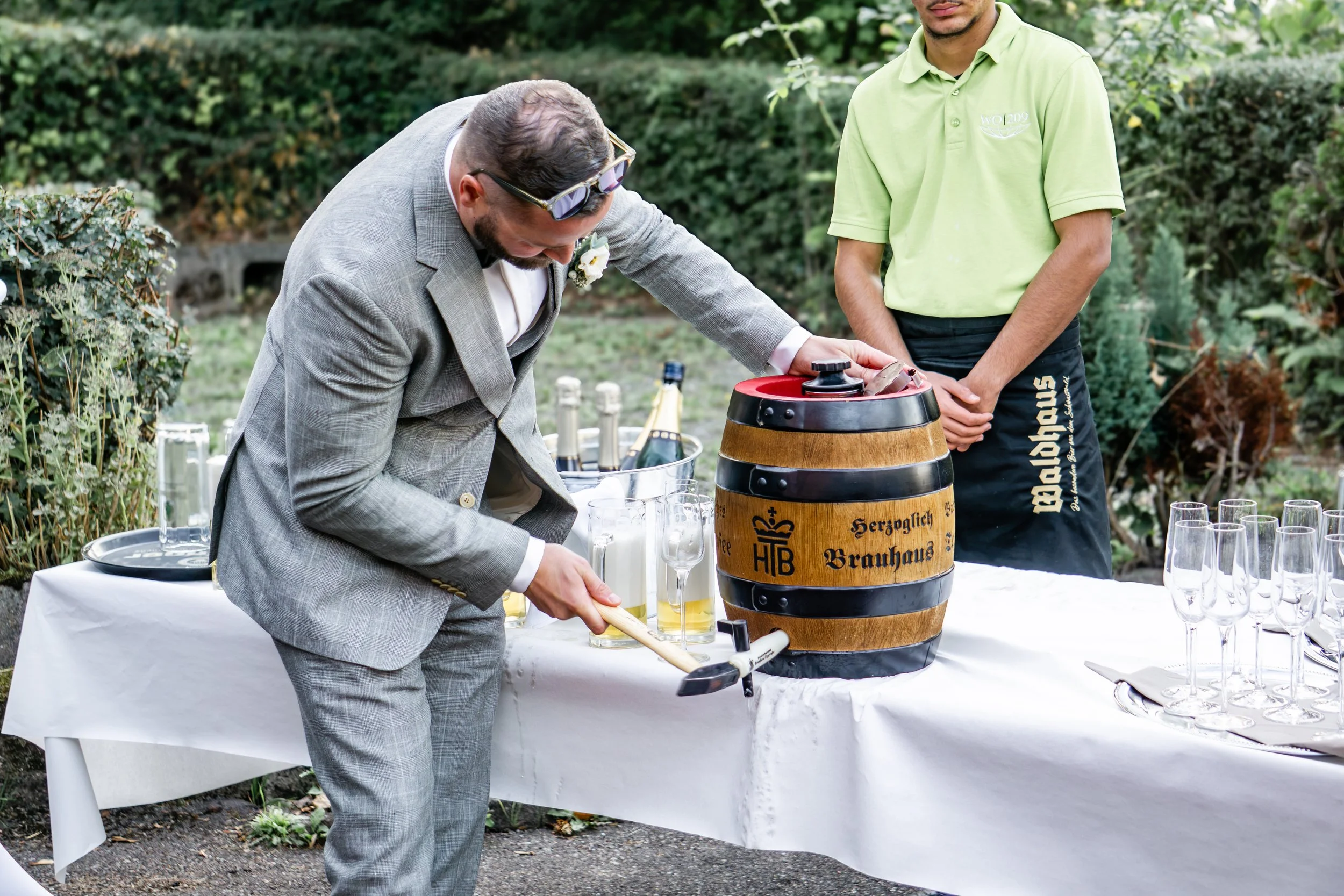 Zwei Männer bei einer Veranstaltung im Freien, einer in einem grauen Anzug, der andere in einem hellgrünen T-Shirt, bereiten Bier aus einem Holzfass auf einer weißen Tafel vor, umgeben von Weingläsern und Flaschen, mit grüner Gartenumgebung im Hintergrund.