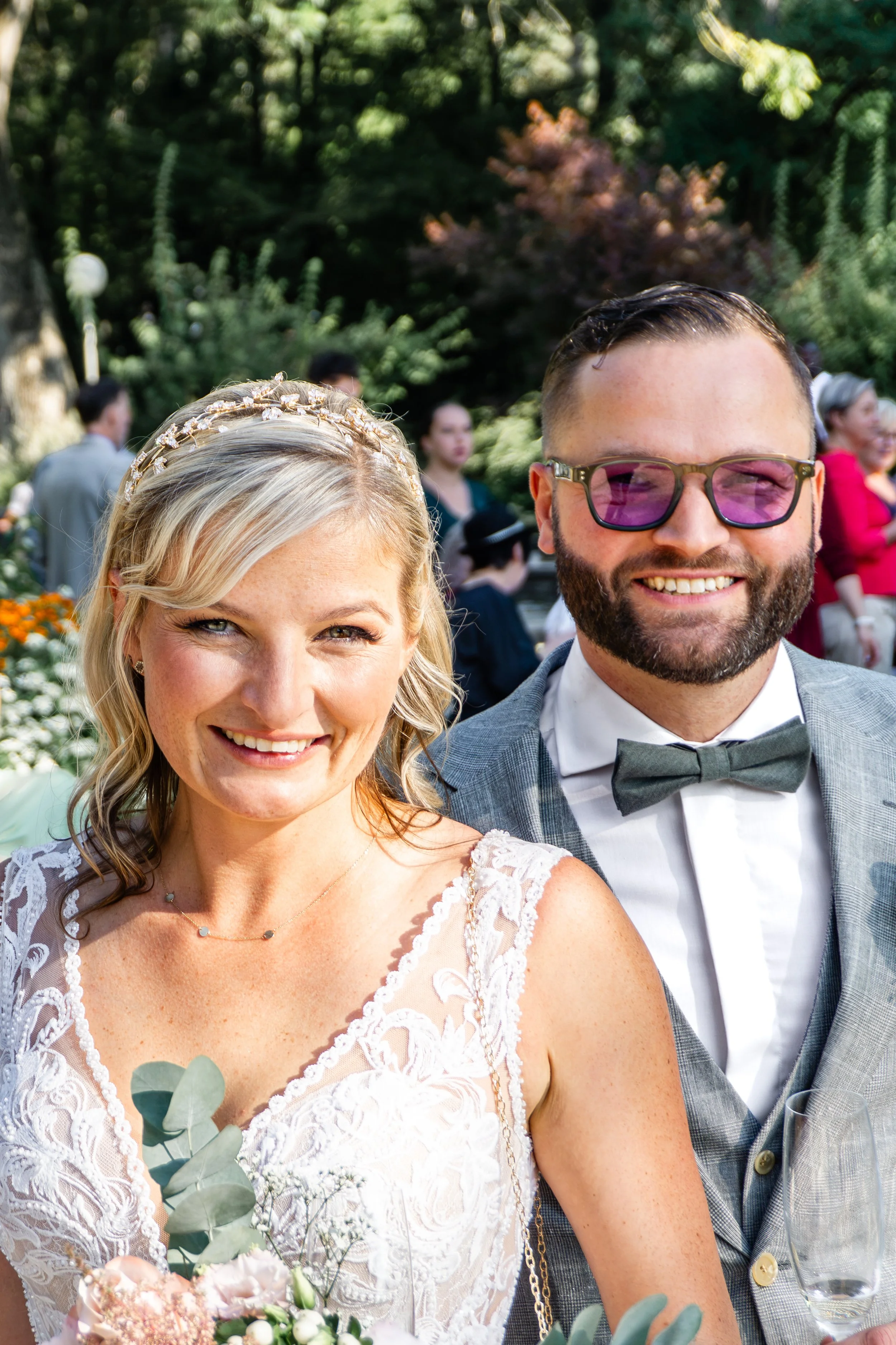 Ein glückliches Brautpaar auf einer Hochzeit im Freien, die Frau trägt ein elegantes Spitzenkleid und die Frisur mit einer goldenen Haarspange, der Mann trägt einen grauen Anzug mit Fliege und Sonnenbrille.