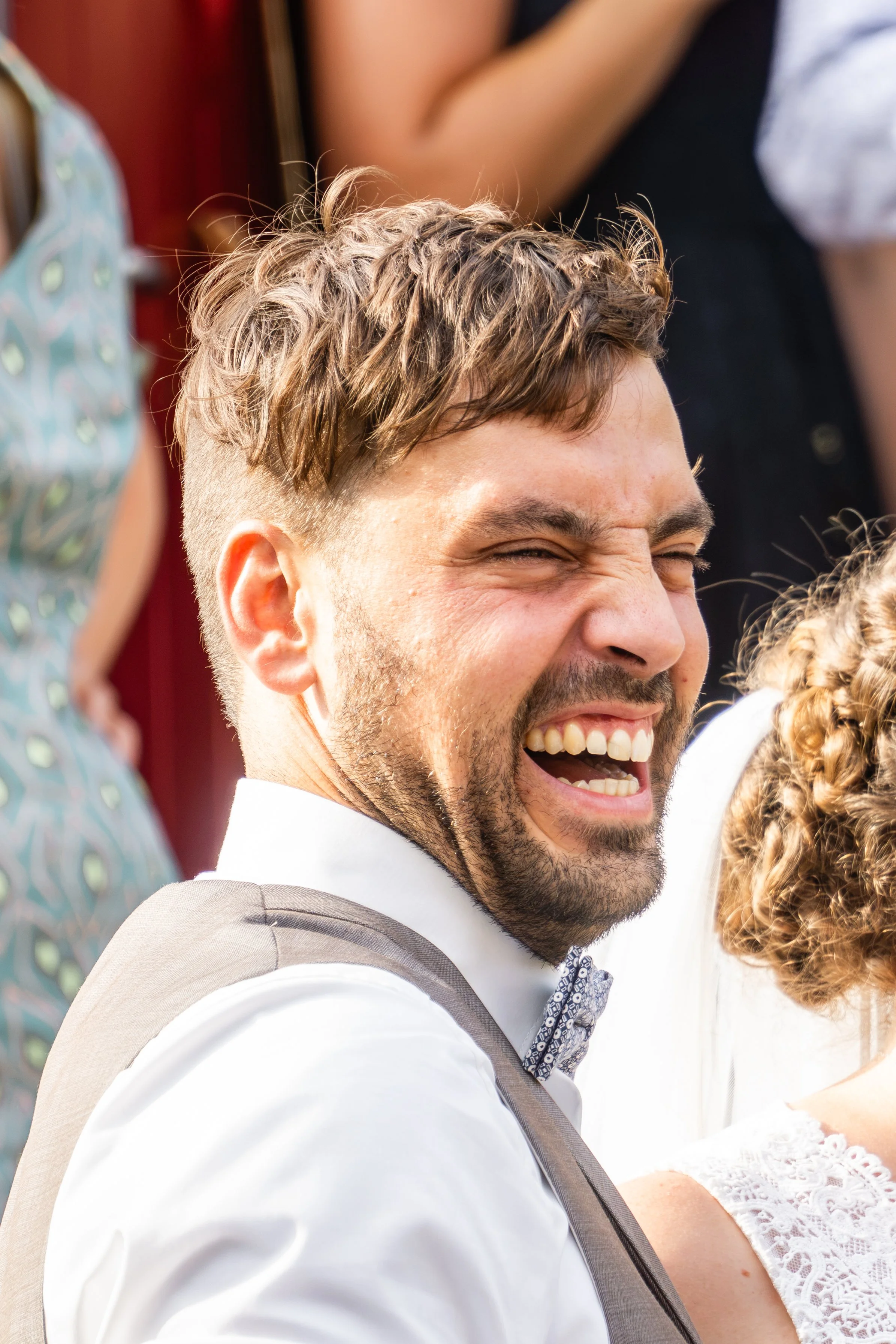 Lachen eines Mannes bei einer Hochzeit in Lübeck, im Hintergrund sind Frauen in sommerlicher Kleidung sichtbar.