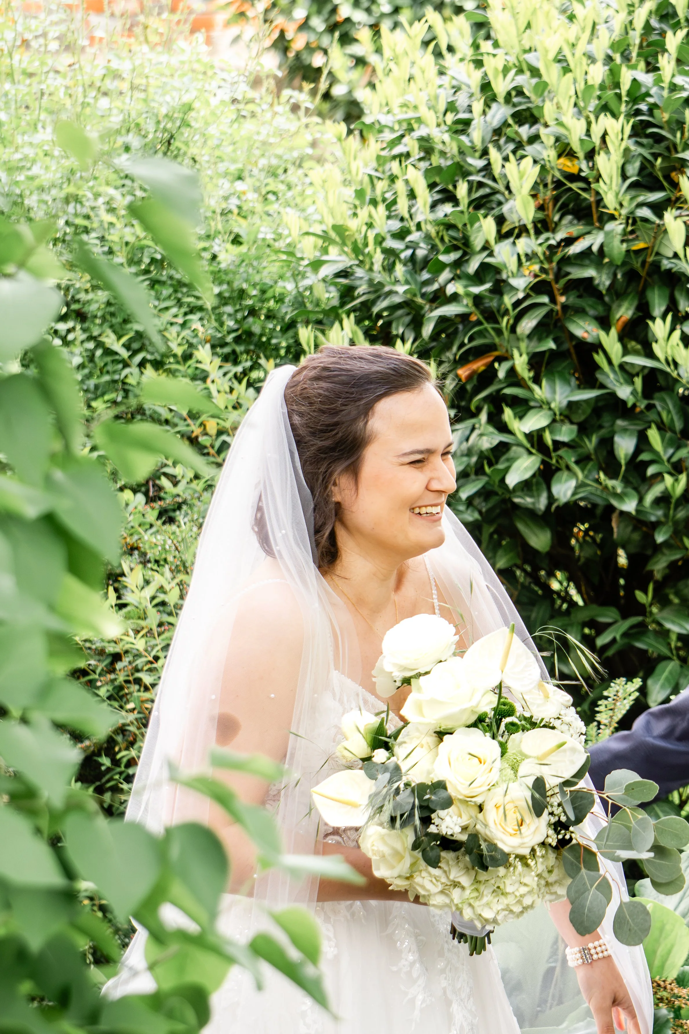 Braut mit dunklem Haar, Brautkleid und Schleier hält einen weißen Blumenstrauß und lacht, umgeben von grünen Blättern.