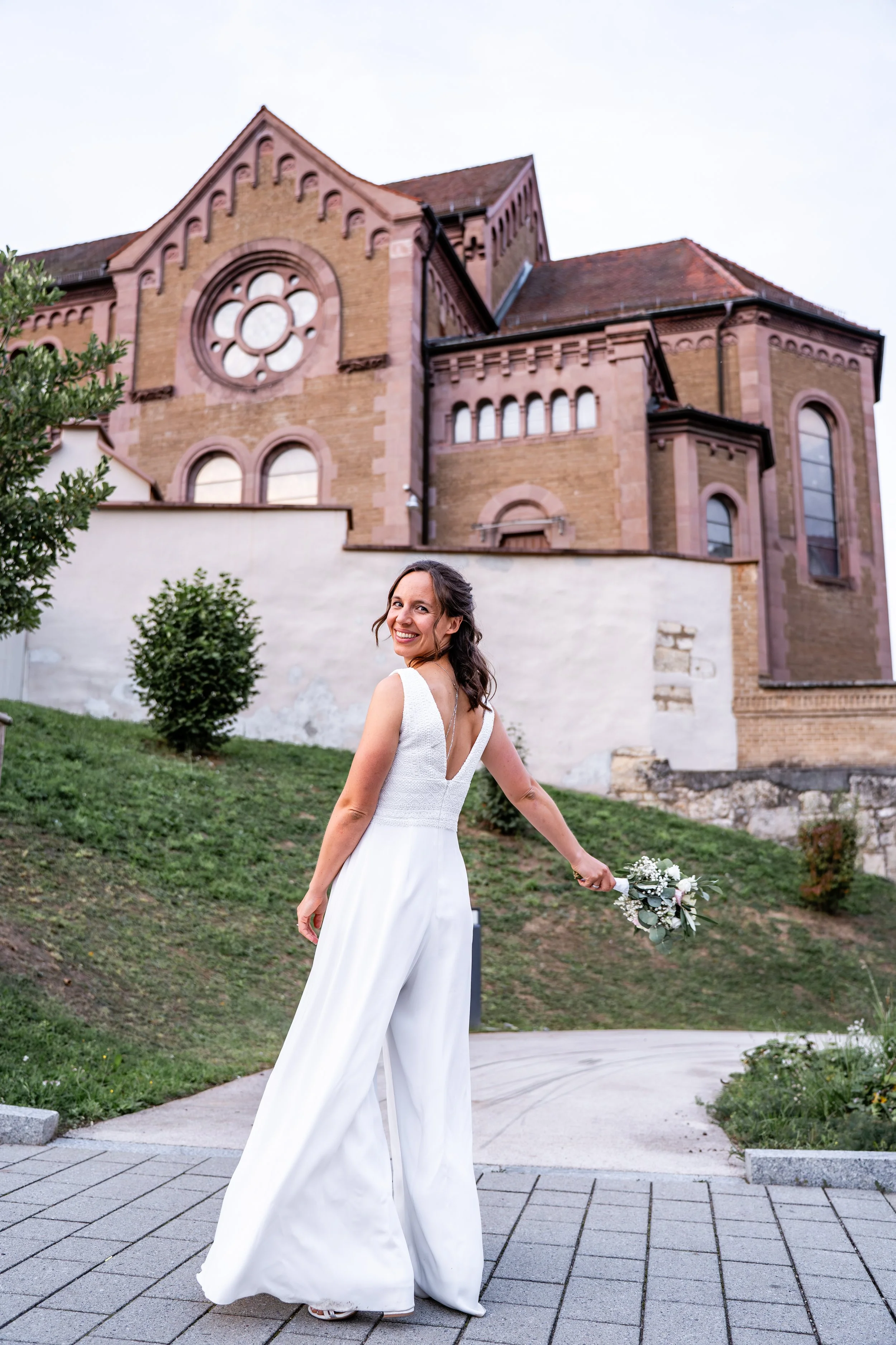 Braut in weißem Kleid hält einen Blumenstrauß, im Hintergrund eine große Kirche aus rotem Stein.