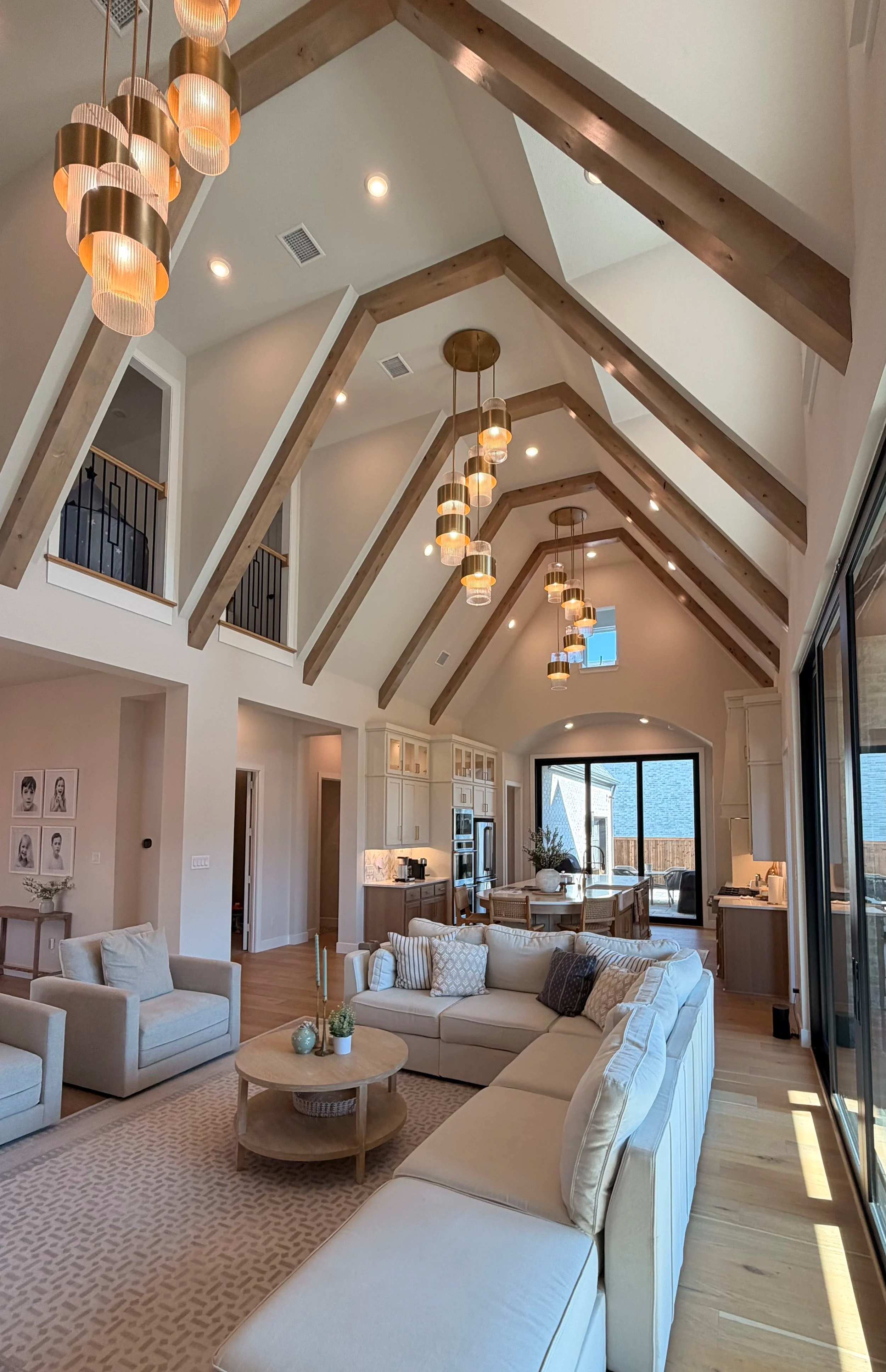 Bright, spacious living room with a high vaulted ceiling supported by wooden beams, modern hanging lights, large windows, and a white sectional sofa with multiple throw pillows.