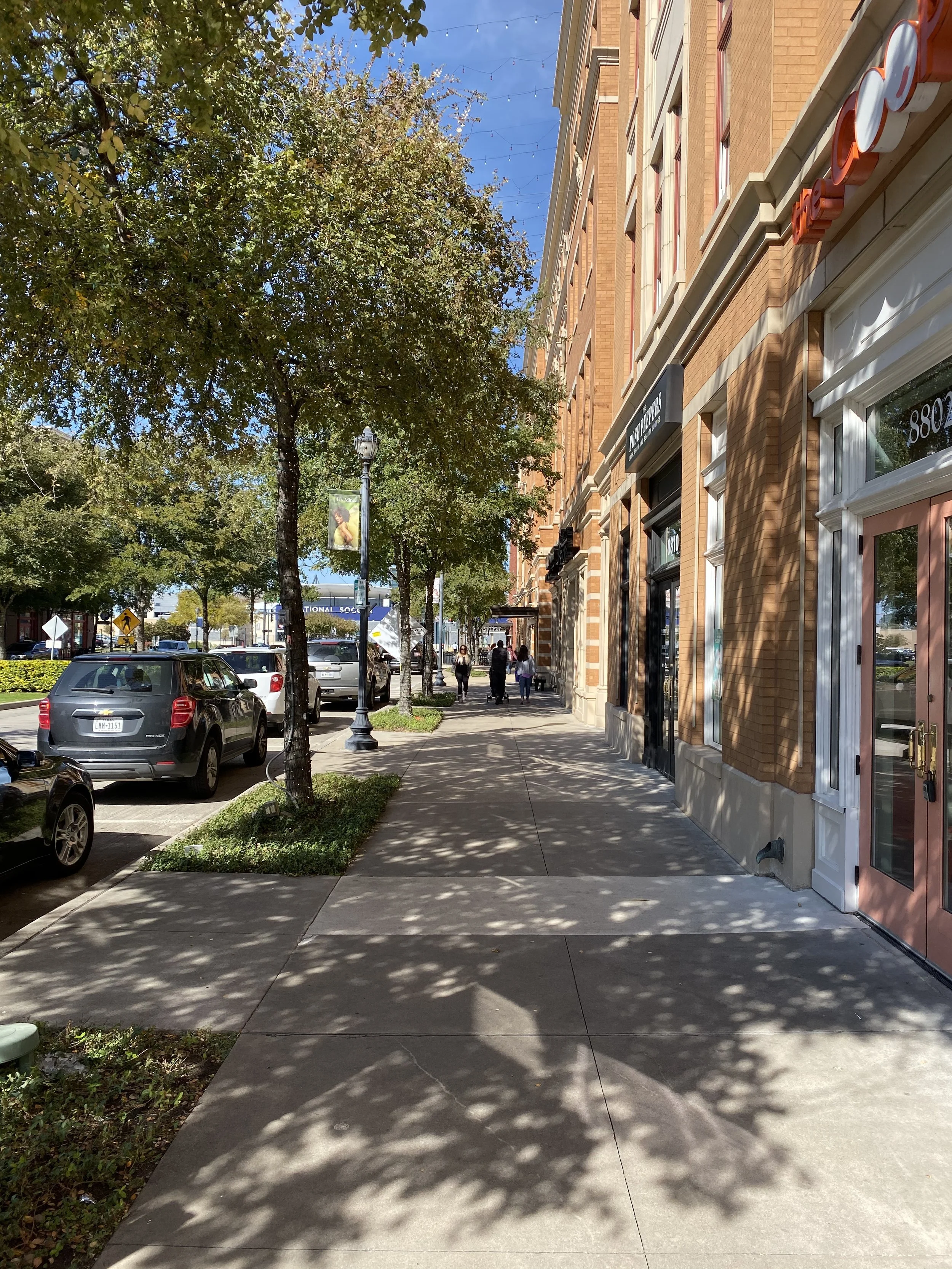 Street view of Frisco Square in Frisco, TX with parked cars, trees, commercial buildings, pedestrians, and a clear blue sky.