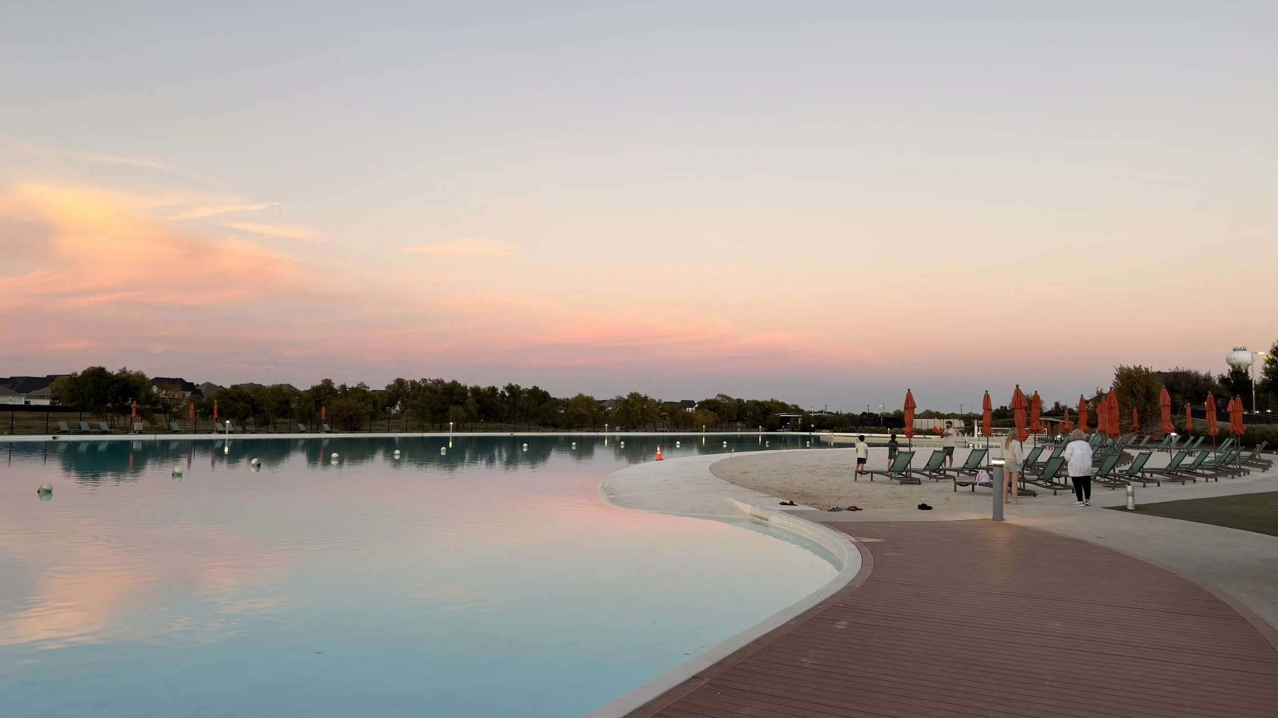 A peaceful outdoor swimming pool scene at sunset at The Lagoon of Windsong Ranch in Prosper, TX with lounge chairs and umbrellas along the poolside, with a calm reflective water and a colorful candy cotton sky.