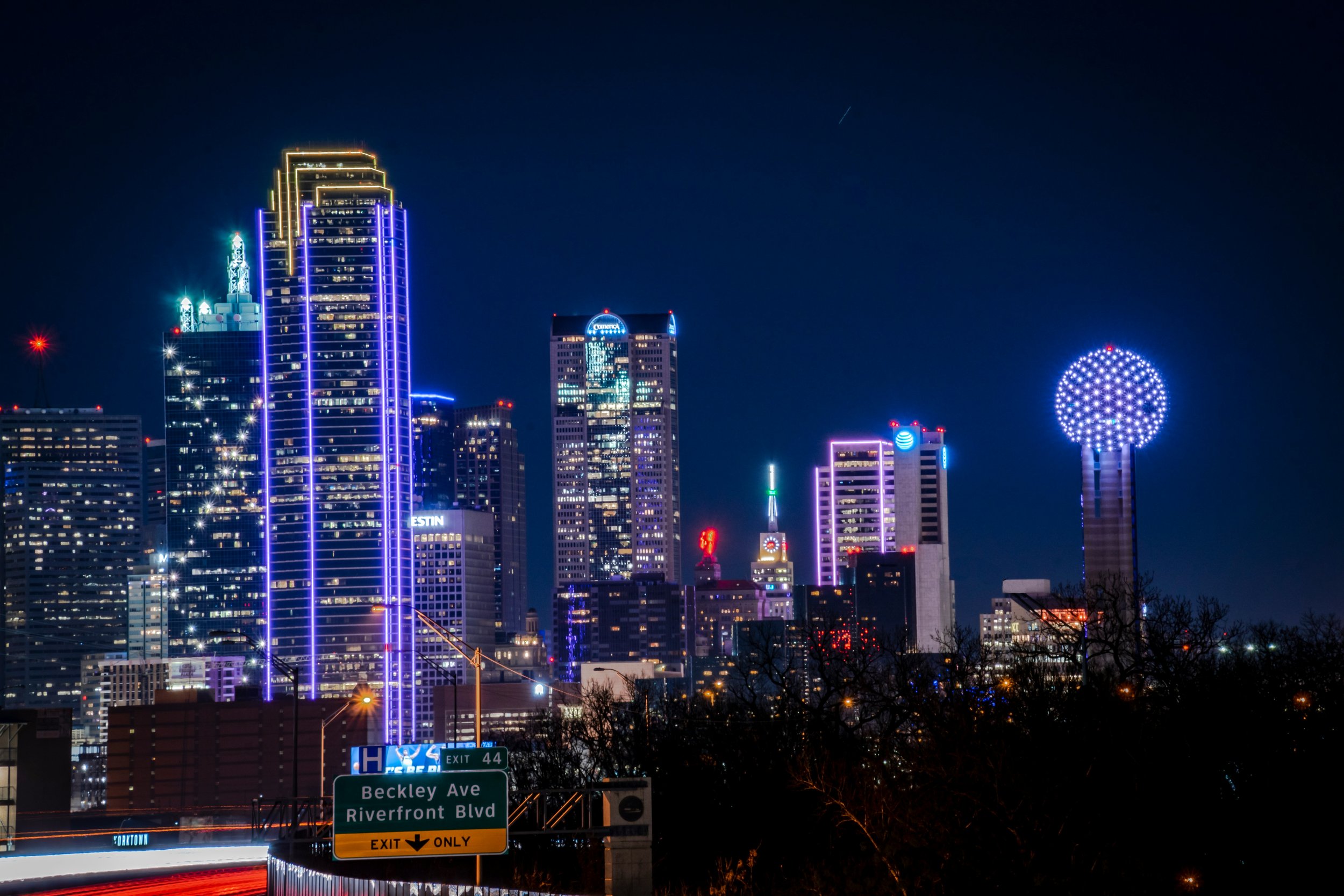 Nighttime city skyline of downtown Dallas, TX with illuminated skyscrapers and buildings, including a tall tower with a spherical top, colorful lights, and street signs in the foreground.