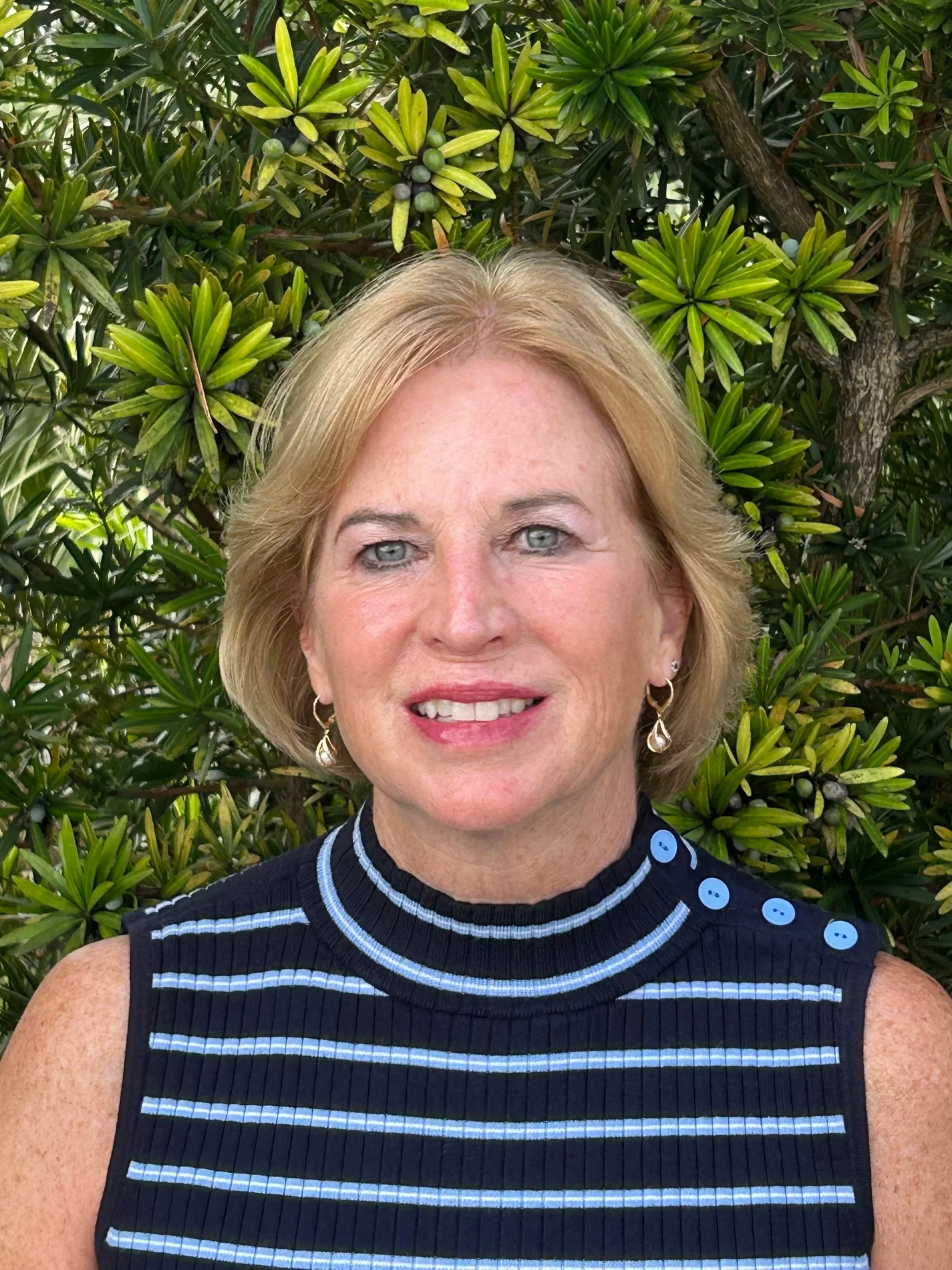 A woman with blonde hair and blue eyes smiling, wearing earrings and a sleeveless navy blue striped top with light blue buttons, standing outdoors in front of green foliage.