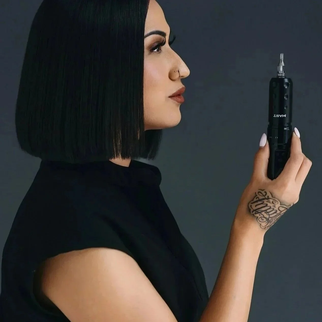 A woman with shoulder-length black hair, light skin, and a nose piercing holding a black tattoo machine against a dark background.