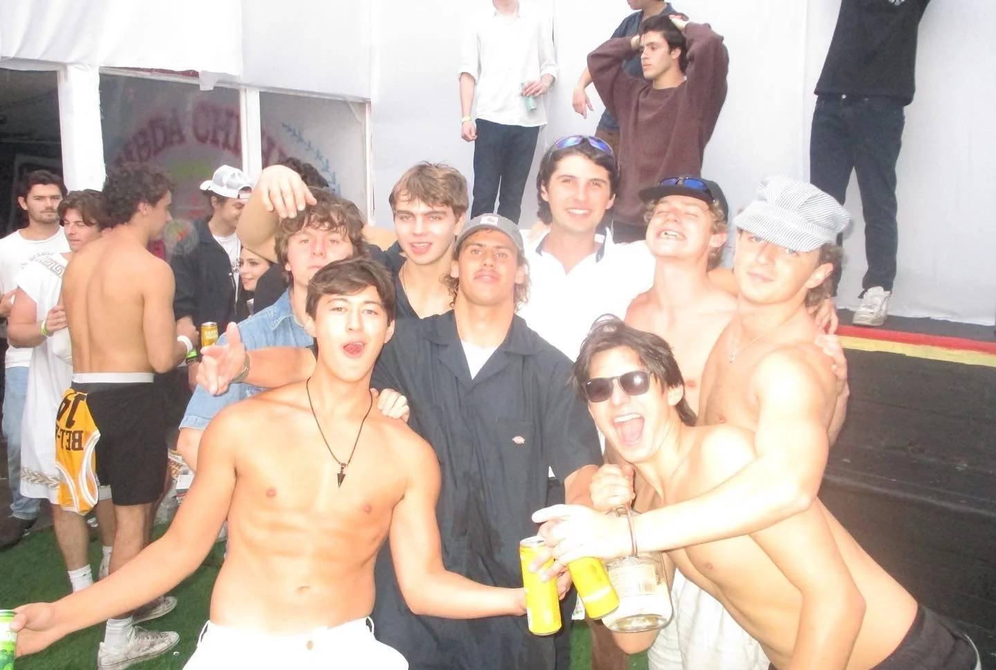 Group of young men at a party, some shirtless, smiling and holding drinks, with a stage in the background.