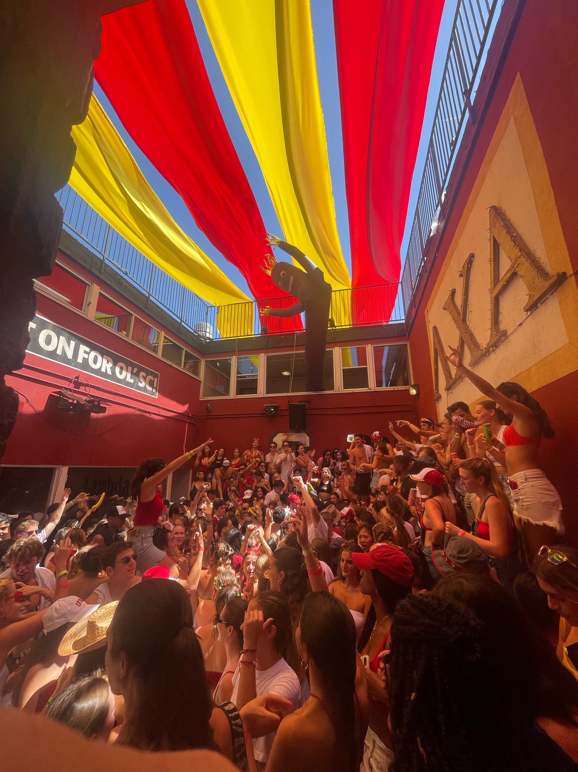 Crowd of people gathered inside a colorful building decorated with red and yellow fabric, with a balcony overlooking the scene and a sign that reads 'CONFORO SIC'.
