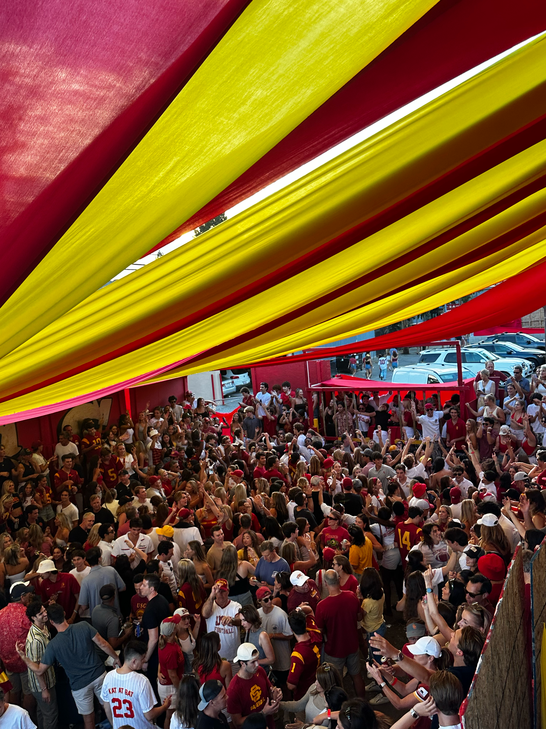 Crowd of people, many in red and yellow clothing, gathered at a sports event under a covered area, with a row of parked cars visible in the background.