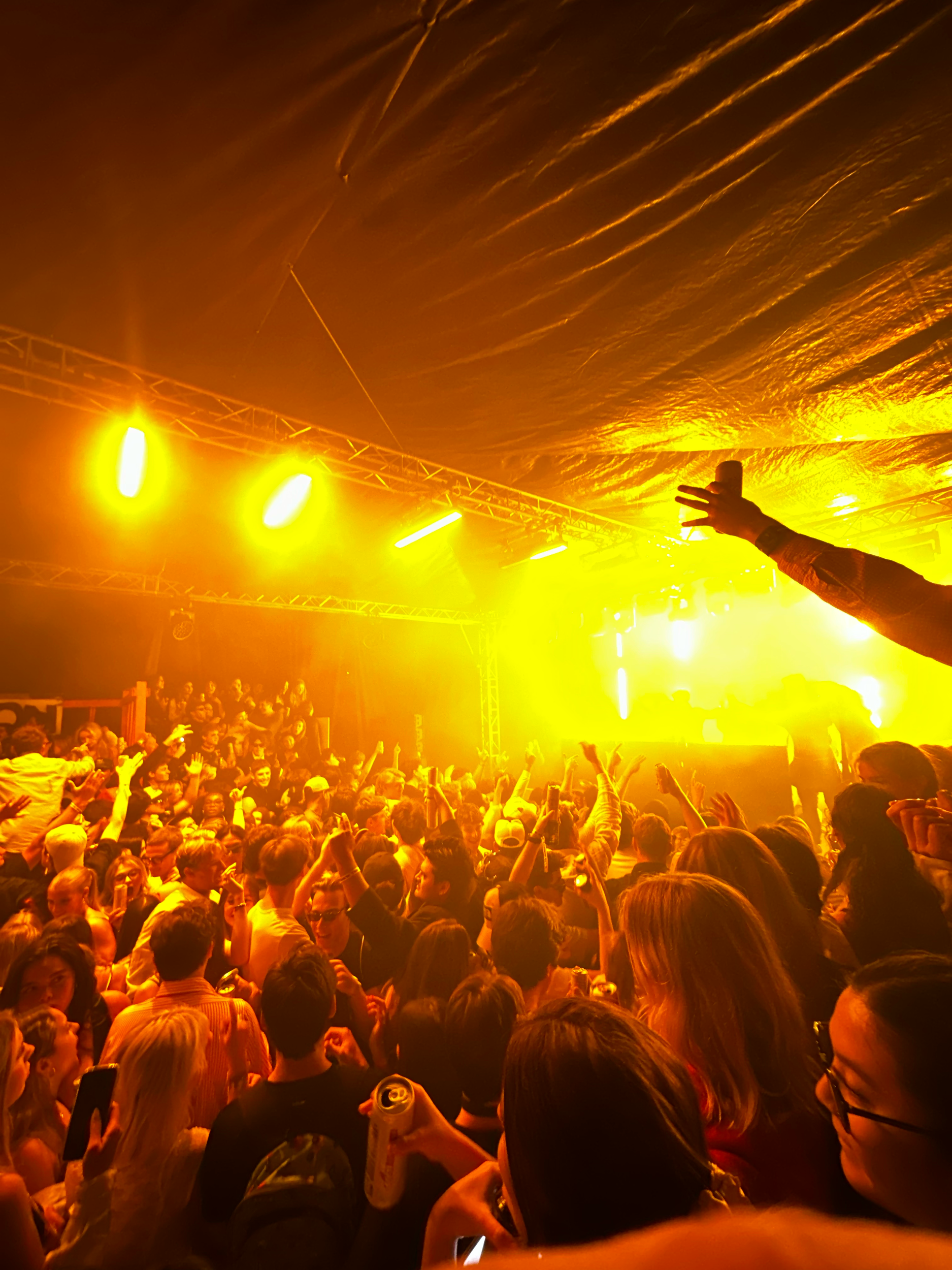 A large crowd of people in a concert or nightclub, with bright yellow and orange stage lights illuminating the scene. The audience is standing close together, some with drinks, enjoying the live performance.
