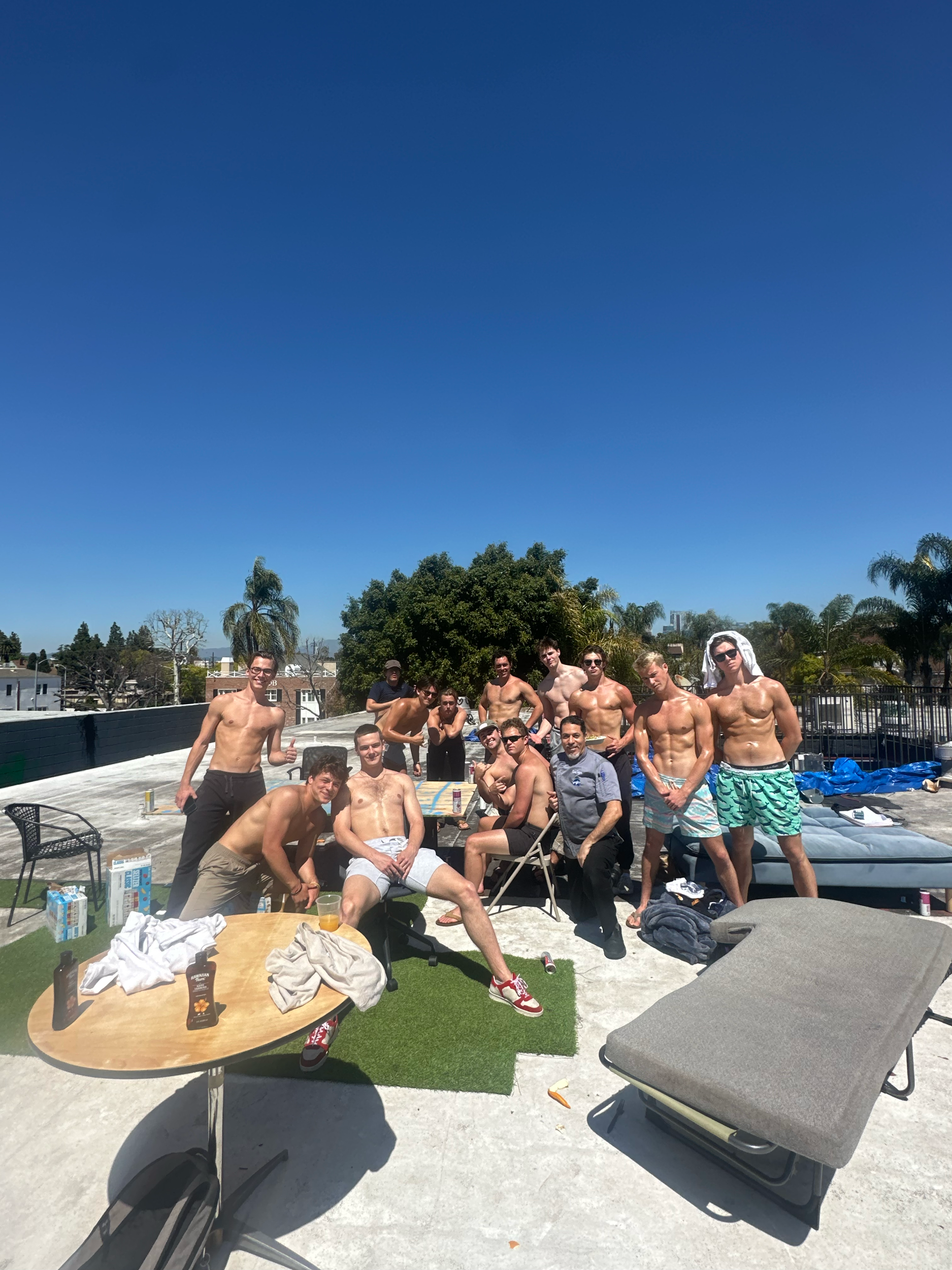 A group of people in swimwear gathered together outdoors on a sunny day, posing for a photo on a concrete platform near a pool, with trees and a clear blue sky in the background.