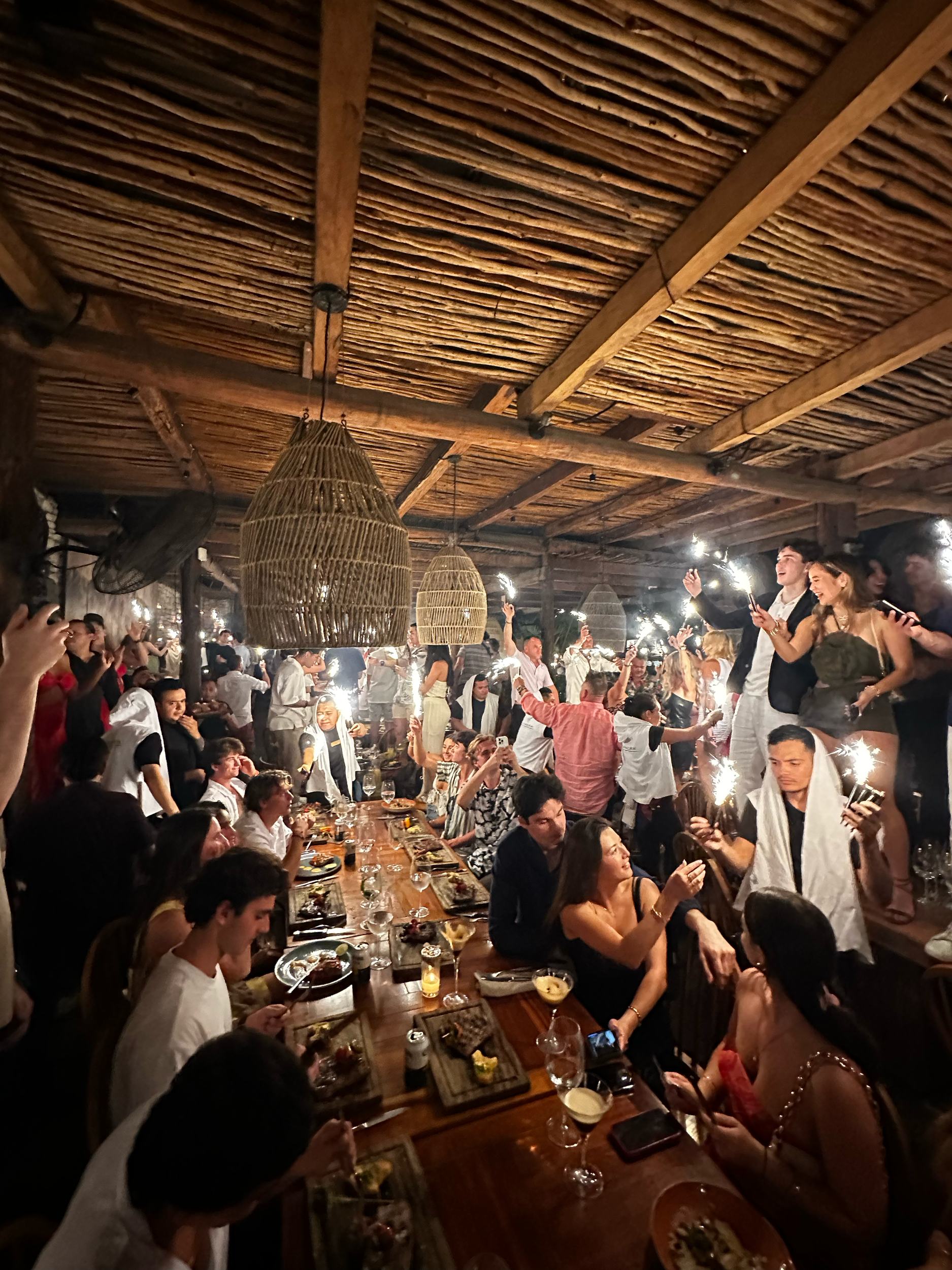 Crowd of people celebrating in a rustic indoor space with bamboo walls, holding sparklers, and gathered around a long dining table with food and drinks.