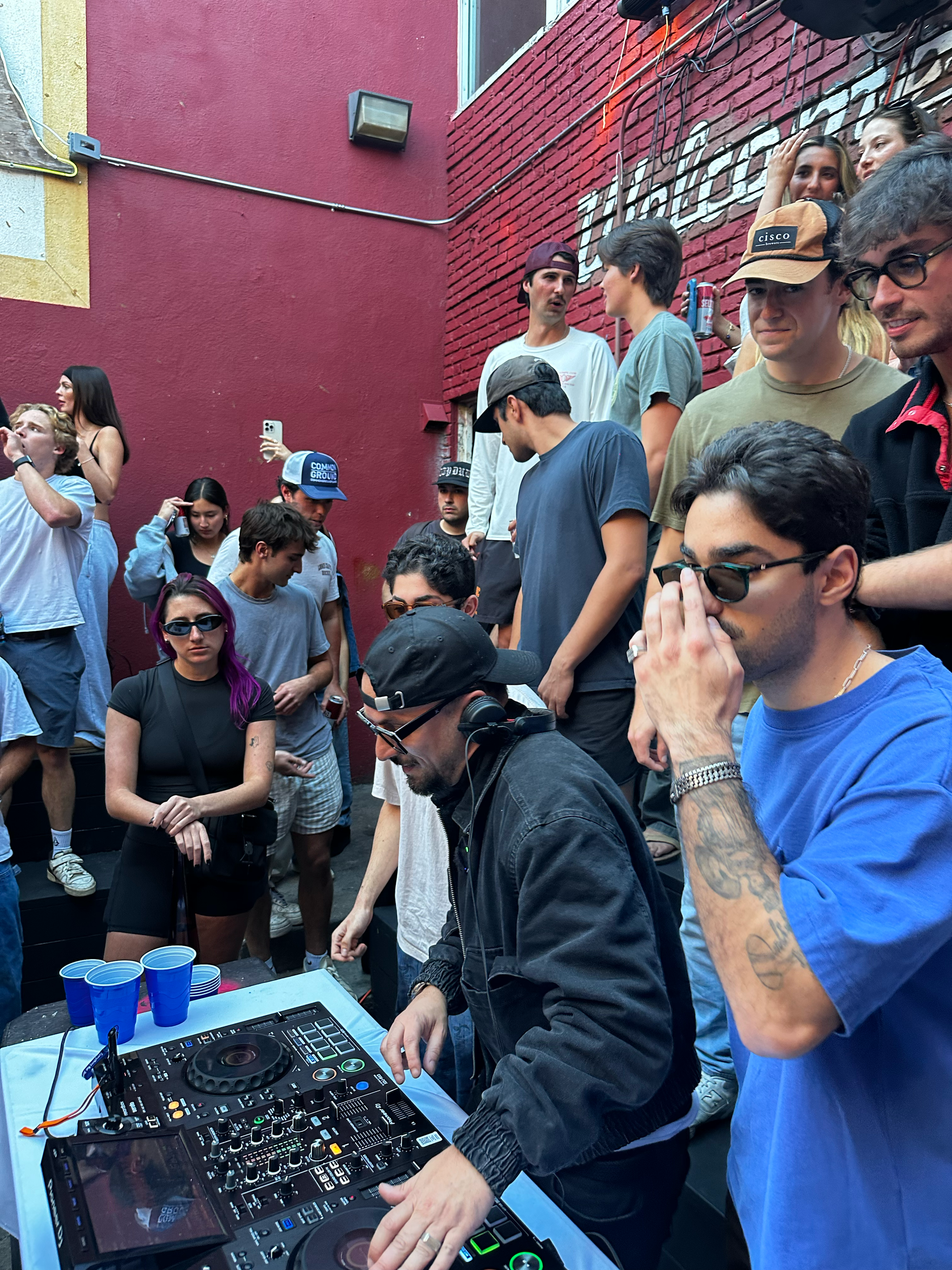 A DJ is playing music at an outdoor event or party, with a crowd of people standing around. The DJ is wearing a black hoodie and headphones, operating a DJ controller, while the crowd watches and socializes against a red brick wall.