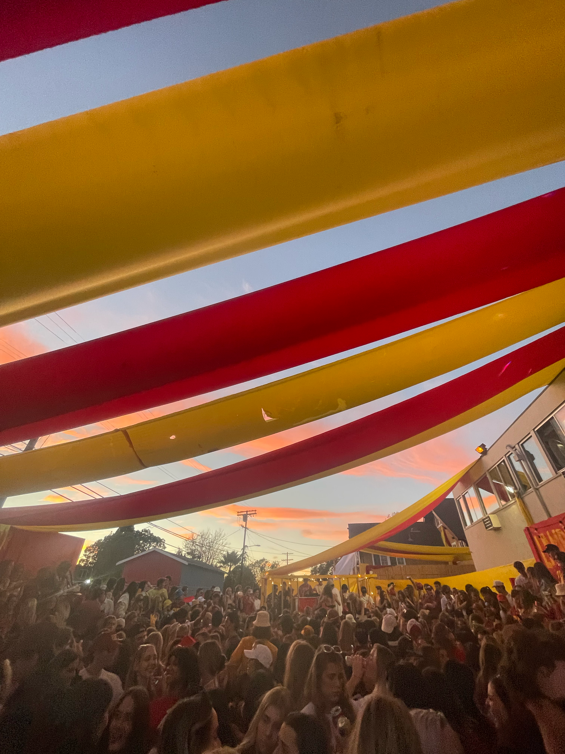 Crowd of people gathered outdoors during sunset, viewed from behind colorful yellow and red railings.