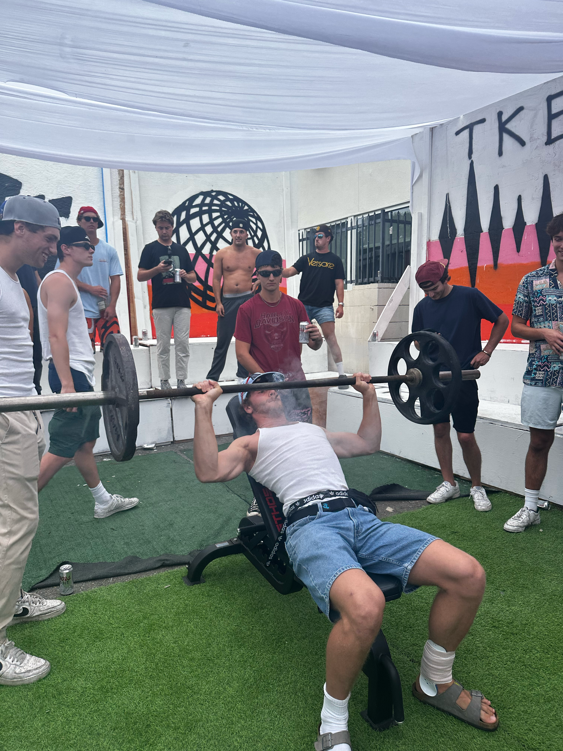 A man performing a flat bench press exercise with a barbell at an outdoor gym. Several people watch, some taking photos. The background features a mural with the word 'TIK' and an abstract globe design.