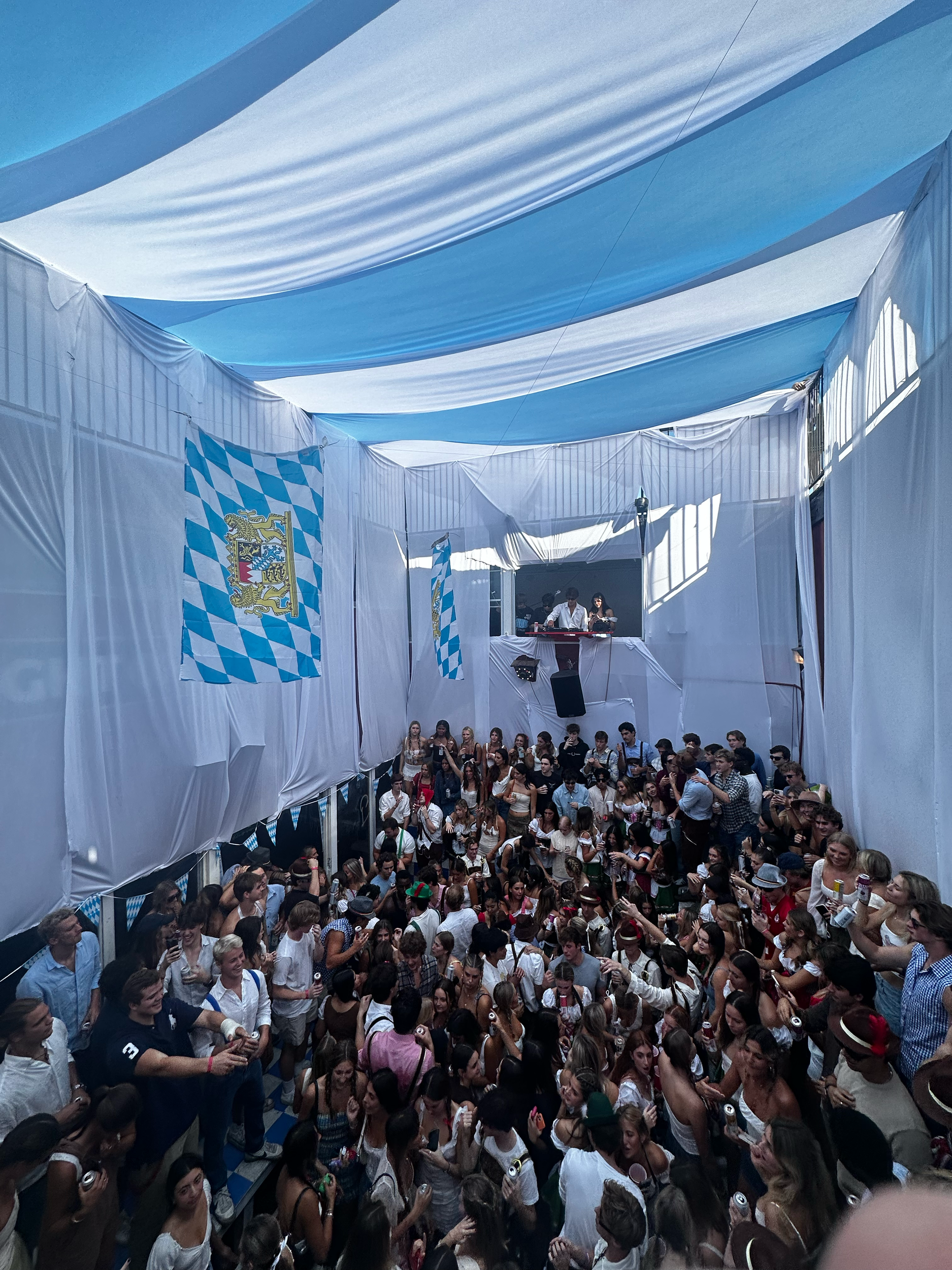 Large crowd gathered at an indoor event decorated with Bavarian flags and decorations, with a stage or DJ booth at the front.