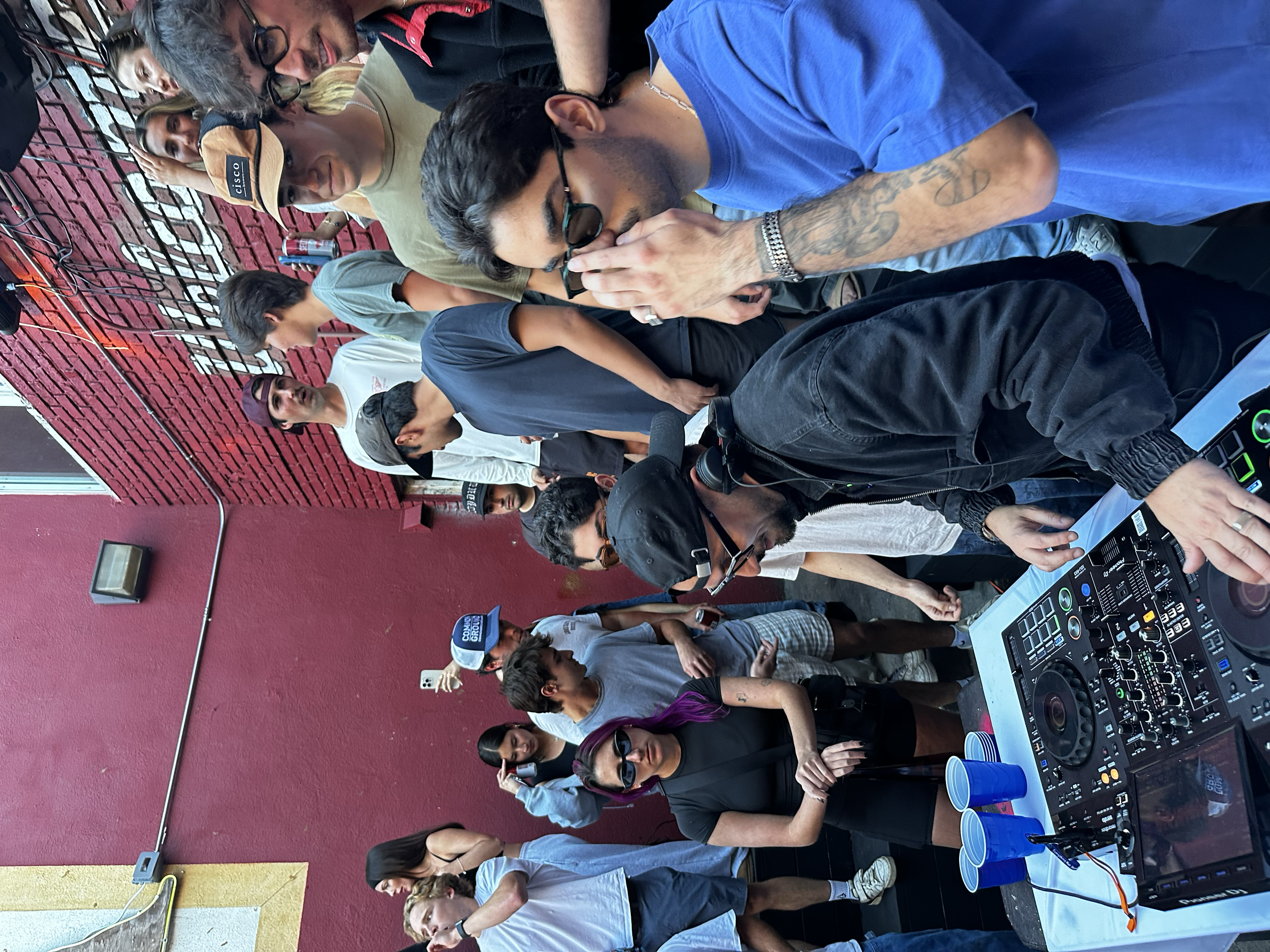A DJ is playing music at an outdoor event or party, with a crowd of people standing around. The DJ is wearing a black hoodie and headphones, operating a DJ controller, while the crowd watches and socializes against a red brick wall.