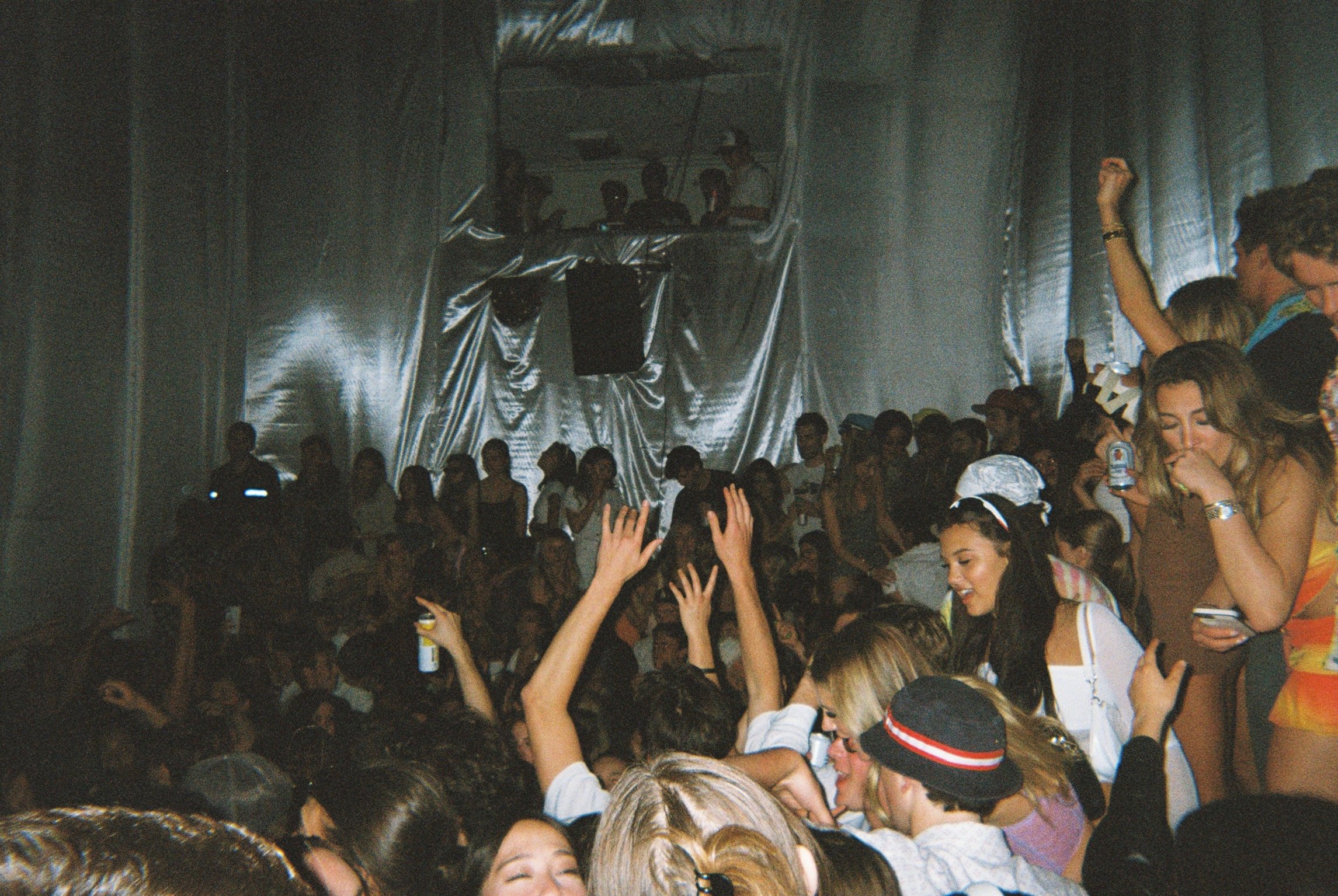 Crowd of people dancing and enjoying themselves at a concert or music event, with a metallic background and a DJ or performer on a raised platform in the distance.