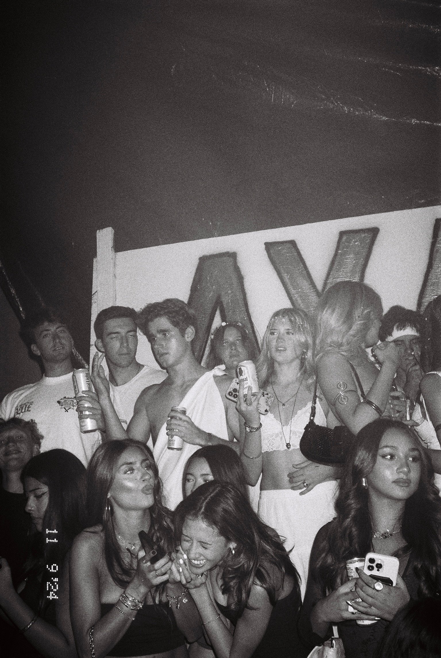 Group of young people at a party, some holding cans of beer, with a large letter sign in the background.